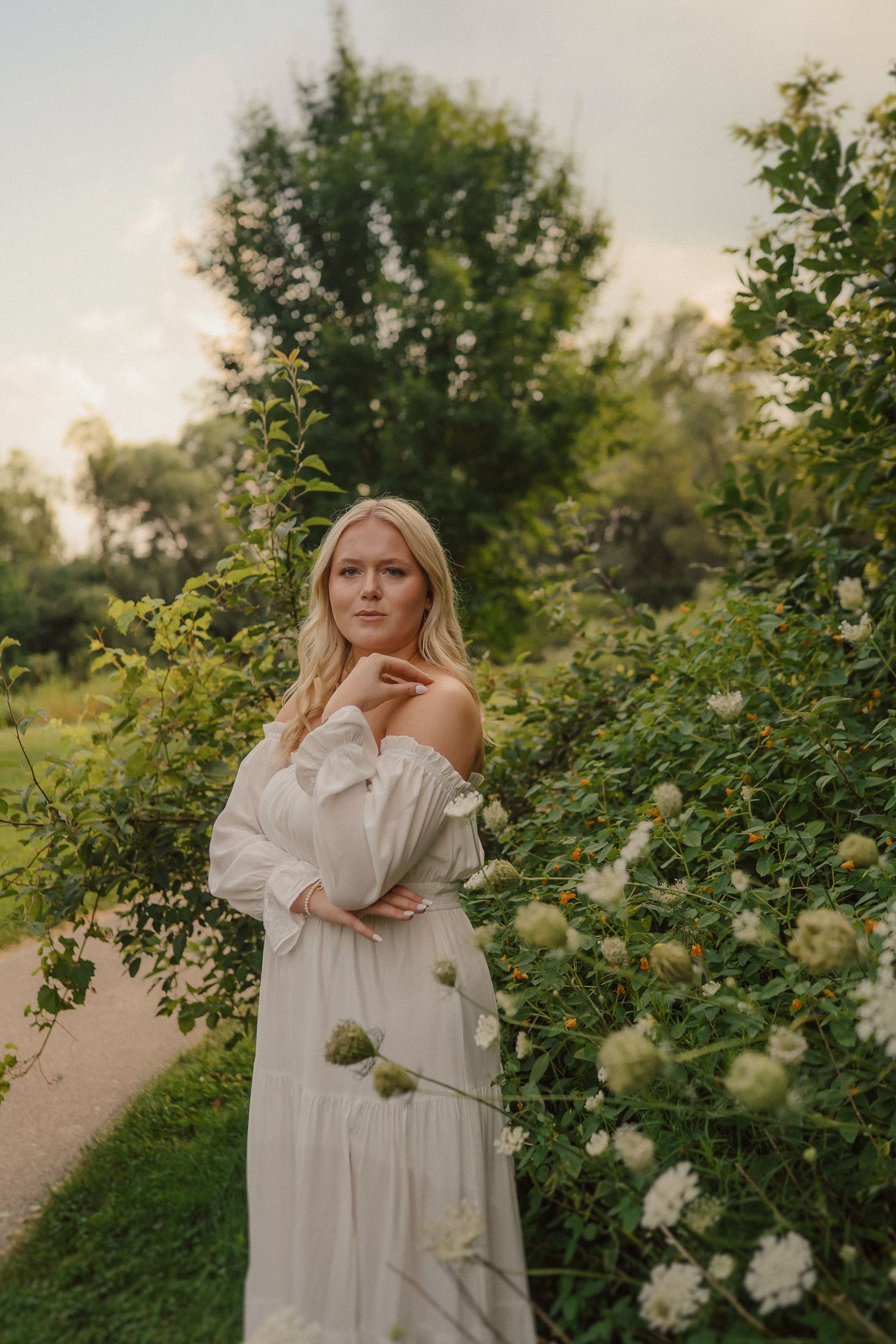 Figure in flowing white dress poses gracefully among blooming white roses along garden pathway at sunset.