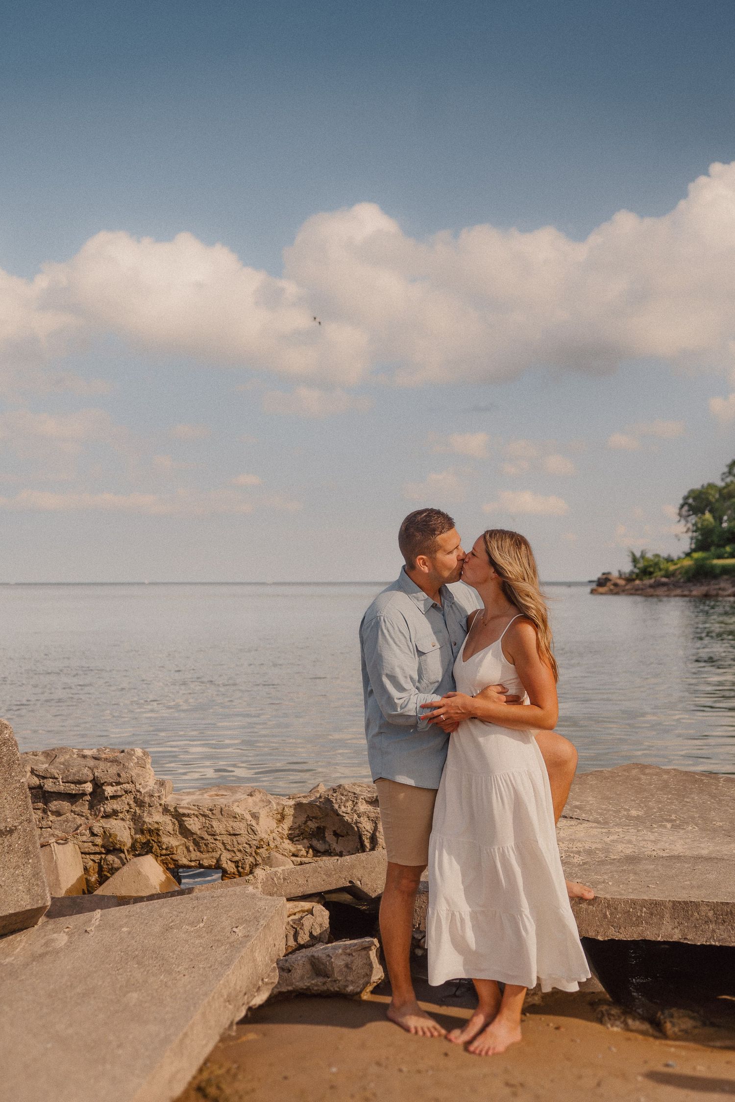 A couple embraces on rocky shoreline during golden hour with calm waters and puffy clouds in background.