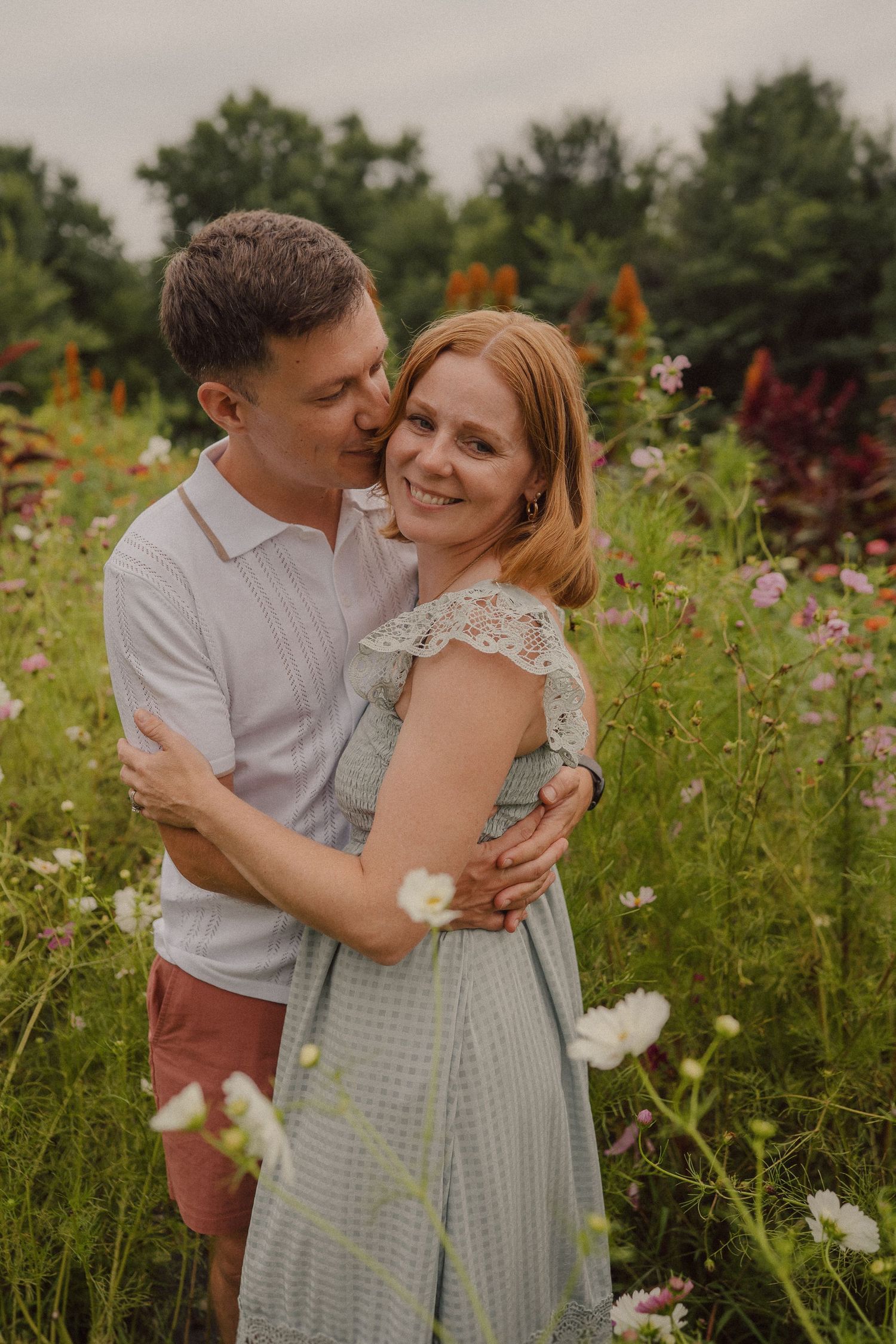 A romantic couple embraces in a green field filled with blooming wildflowers on a warm summer day.