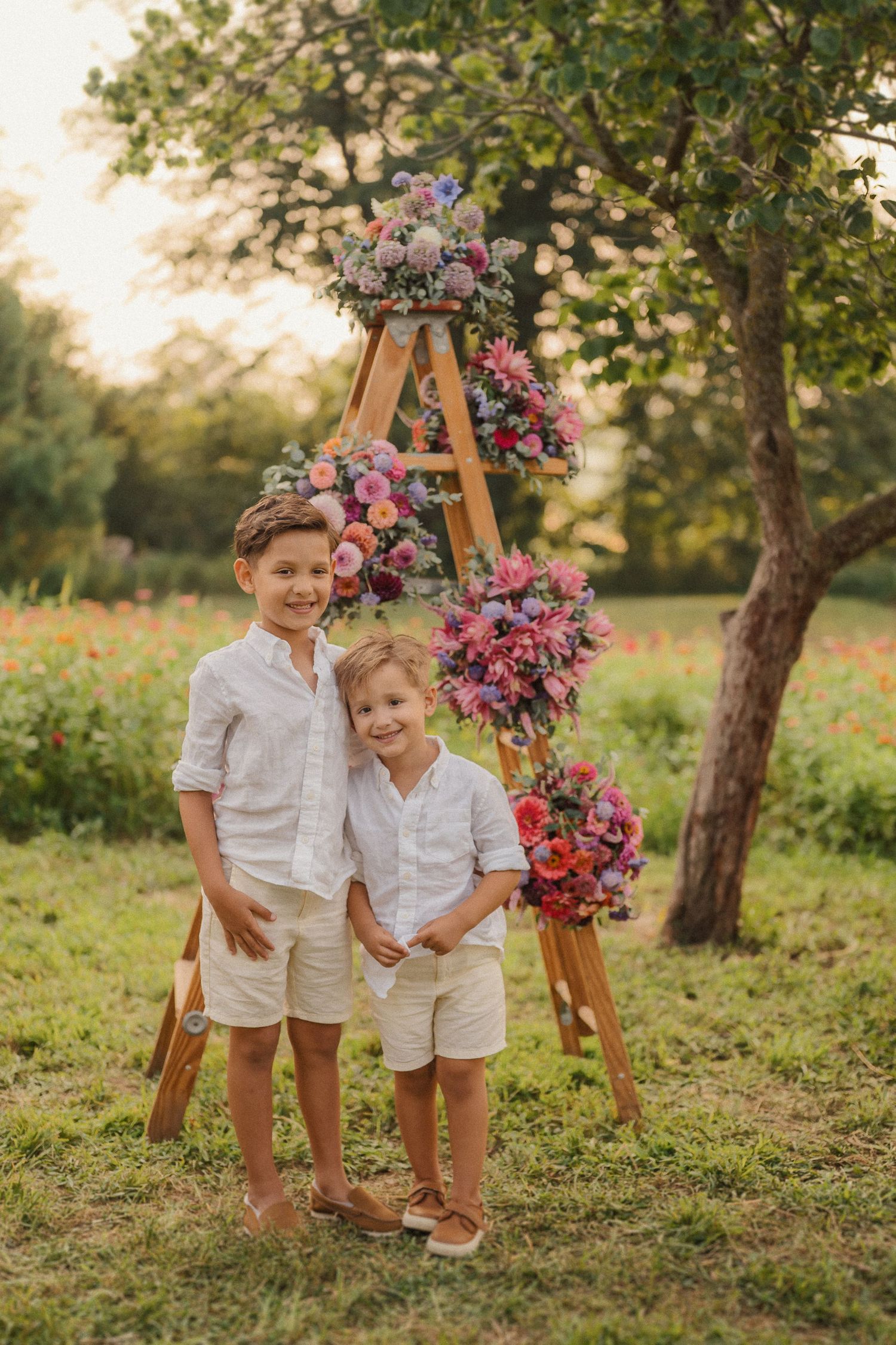 Two children in white shirts and khaki shorts pose in front of a wooden ladder decorated with pink and purple flowers.