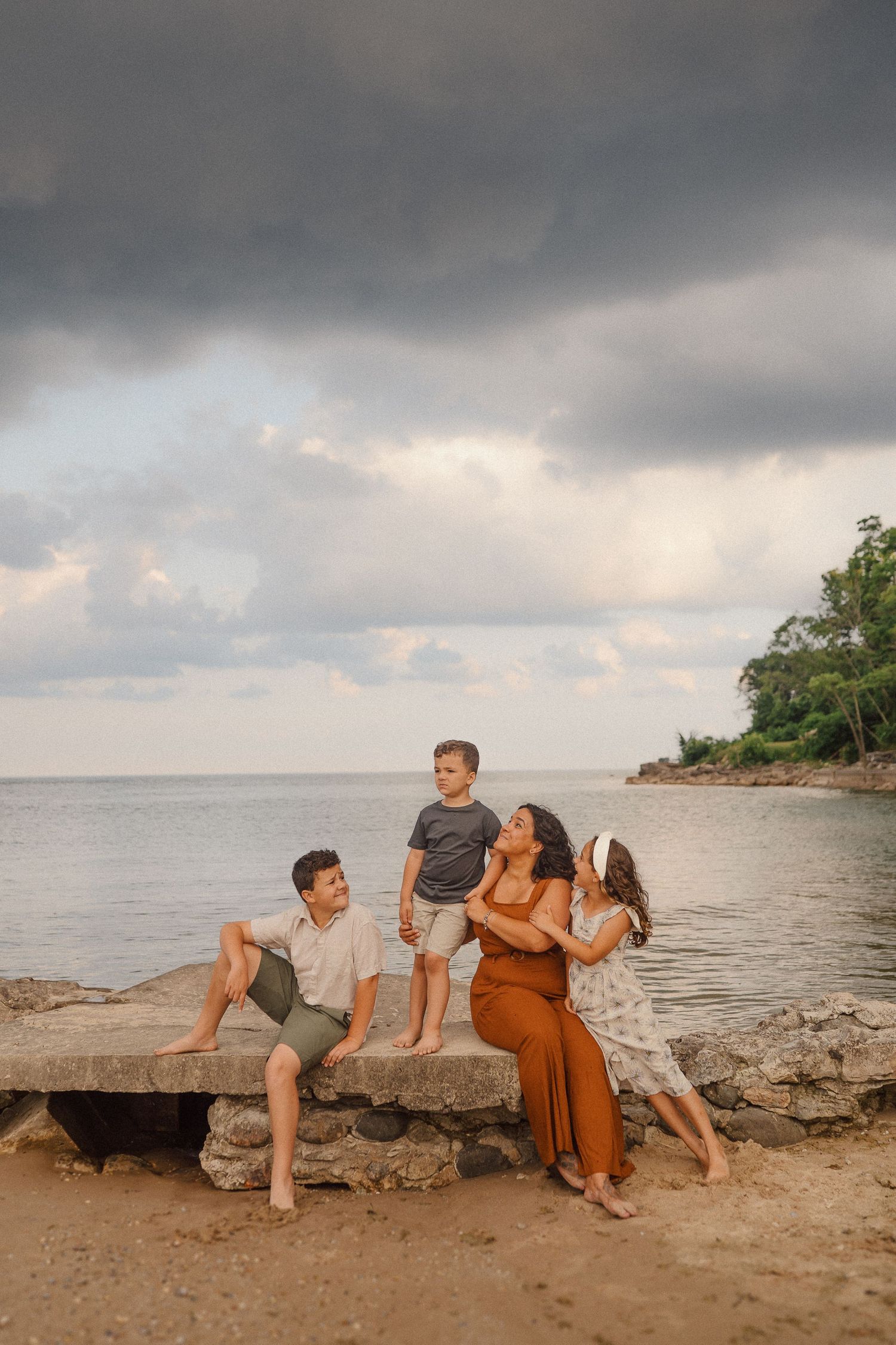 A family enjoys a sunset moment together on rocky beach terrain with dramatic cloudy skies in the background.
