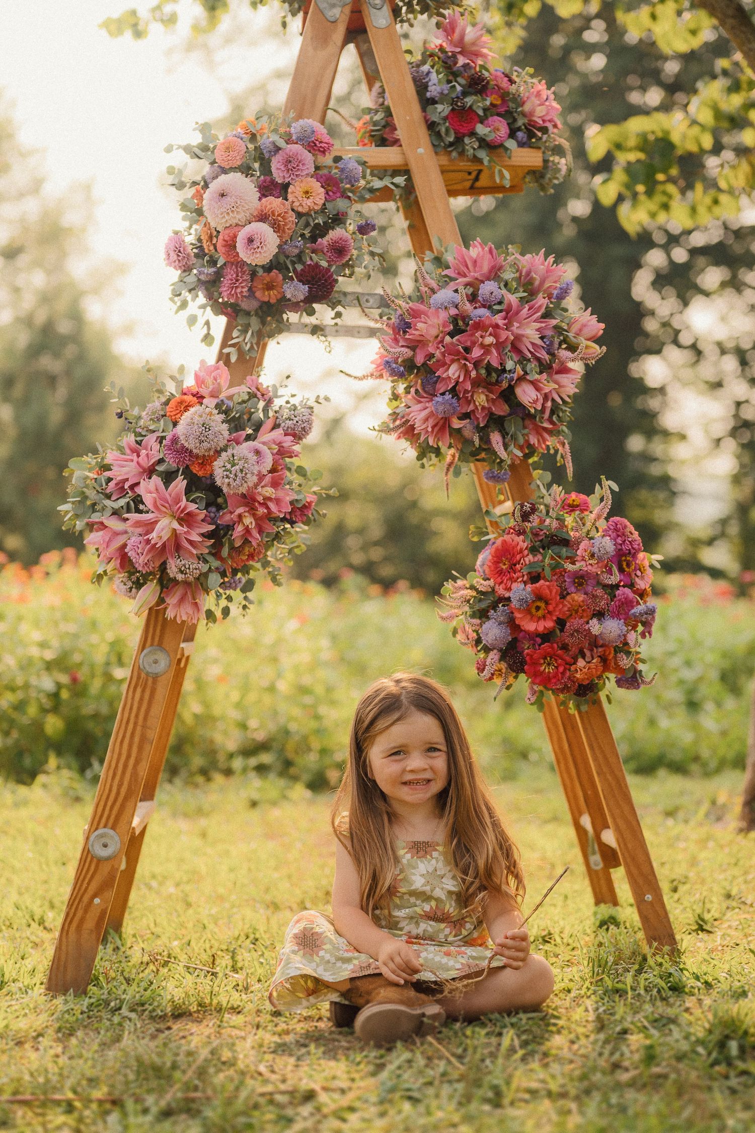 Young child sitting beneath wooden ladder decorated with pink and red floral arrangements in garden.
