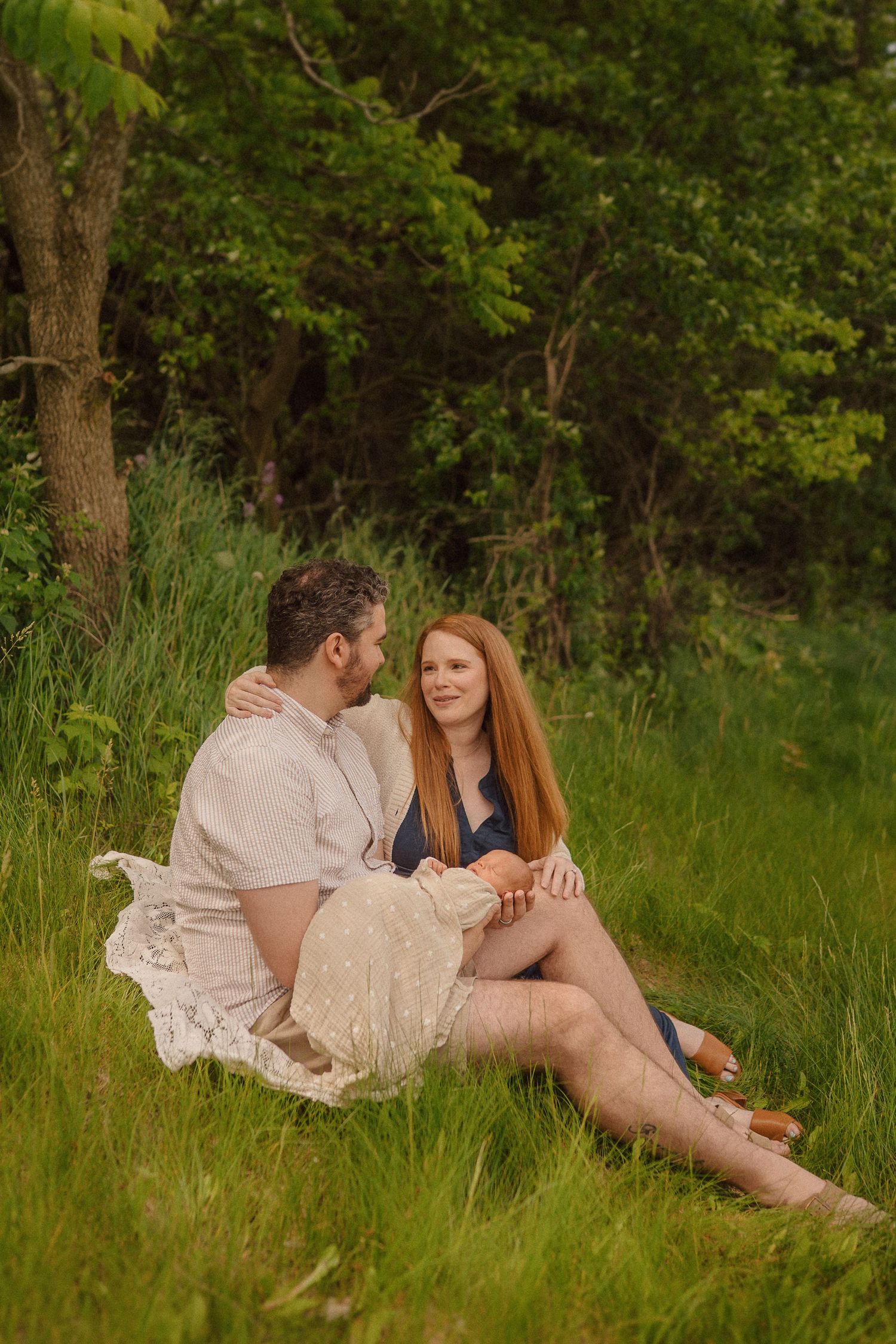 A couple in light summer outfits relaxes together on lush green grass near trees in a romantic outdoor setting.
