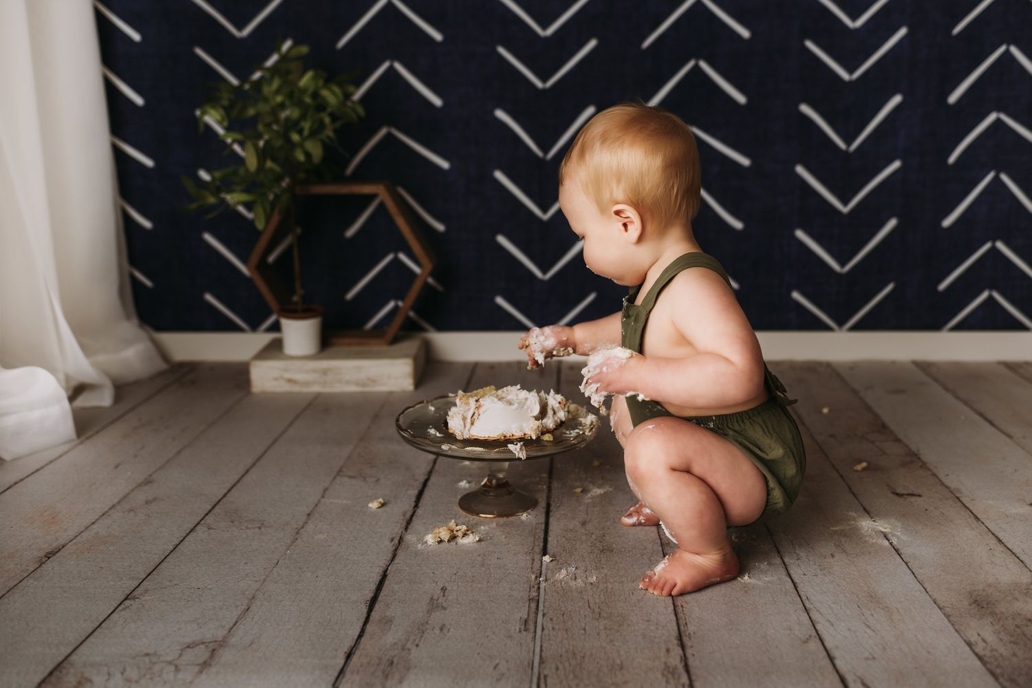 Small child in green outfit explores cake while sitting on rustic wooden floor.