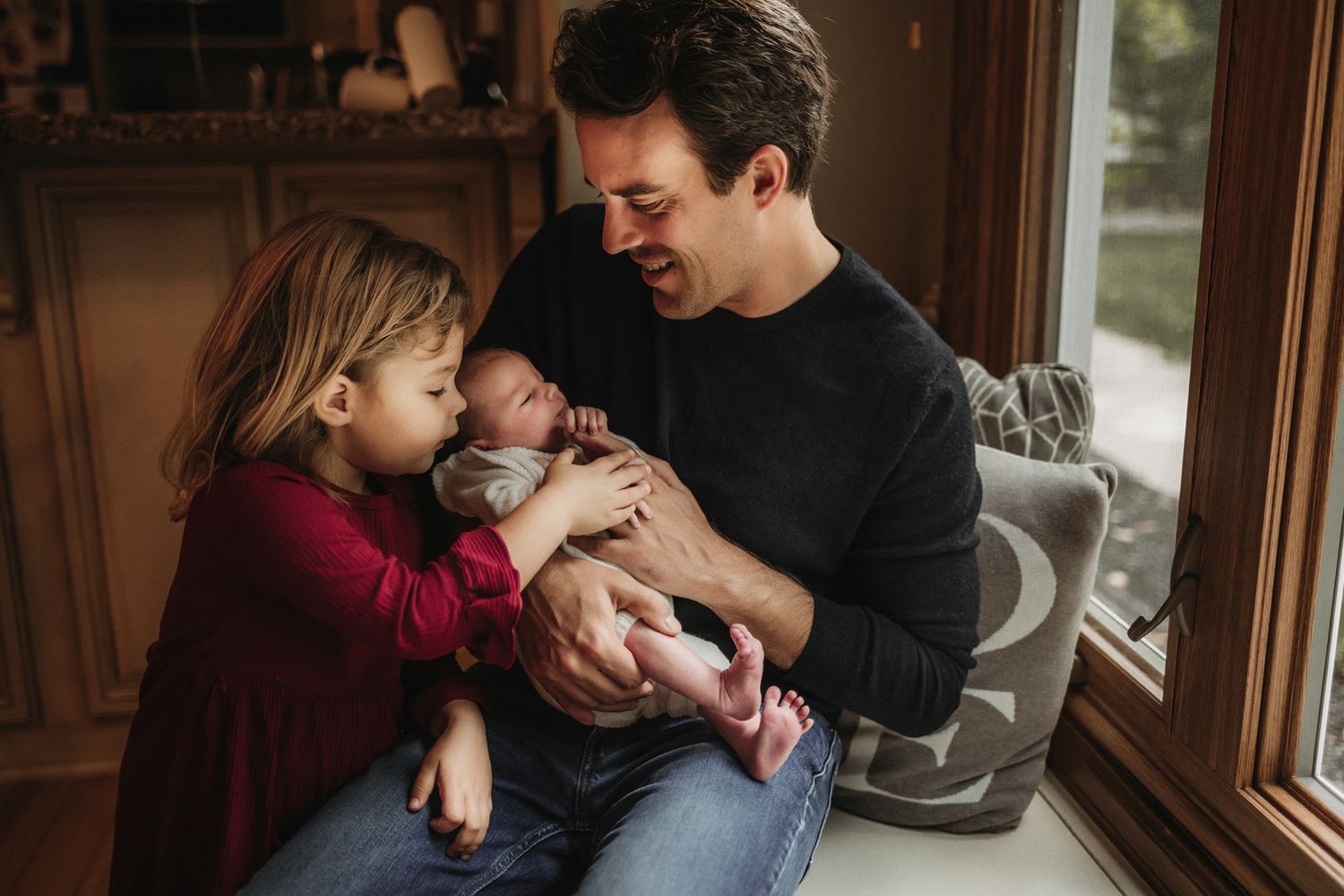 Sweet family moment shared by window with toddler in red dress meeting new sibling.