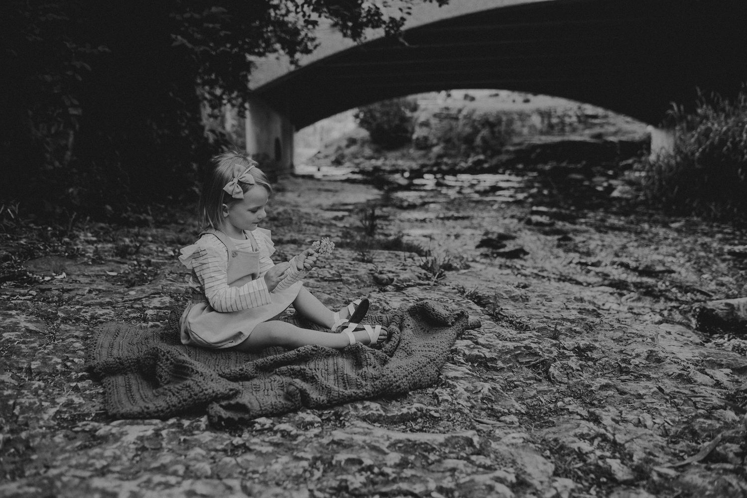 Black and white image of a child sitting on a blanket under a bridge.