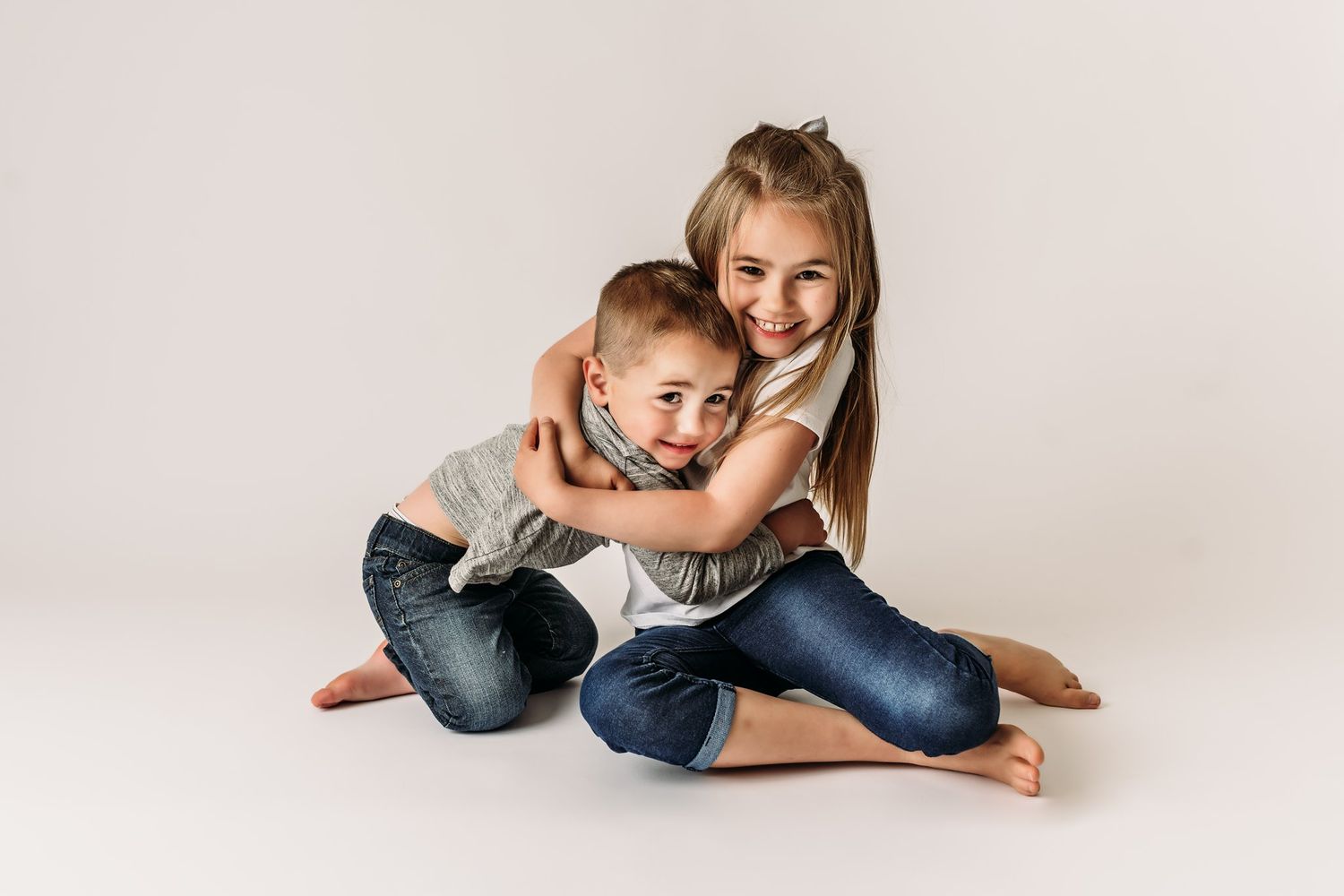 Two siblings embrace while sitting on white studio background.