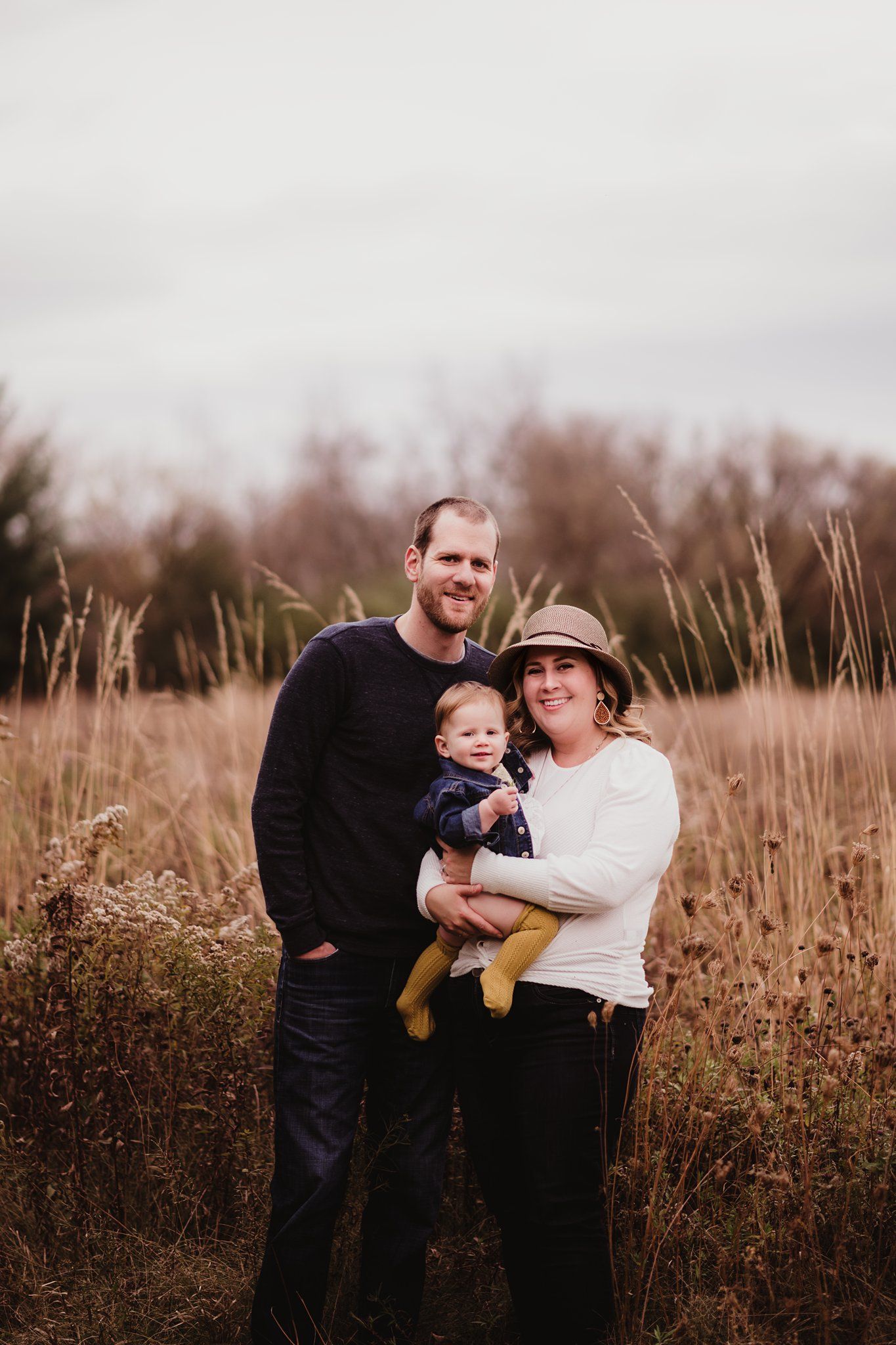 A family of three poses together in a field of tall autumn grass at sunset.