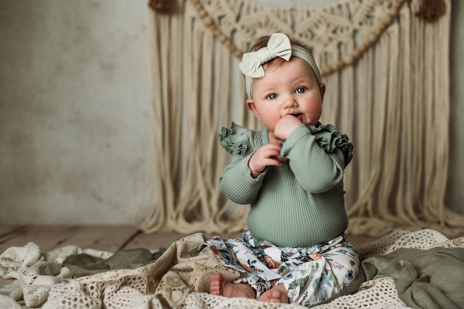 Baby in mint green outfit brings hands to mouth while sitting on cream lace blanket.
