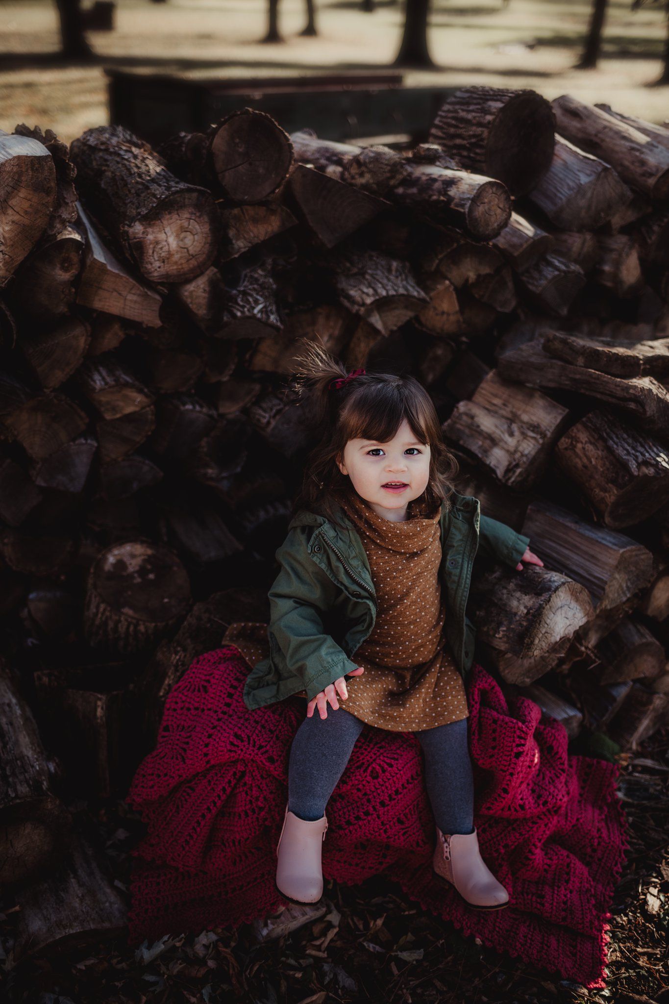 A child in a green jacket sits on a red blanket against a woodpile during autumn.