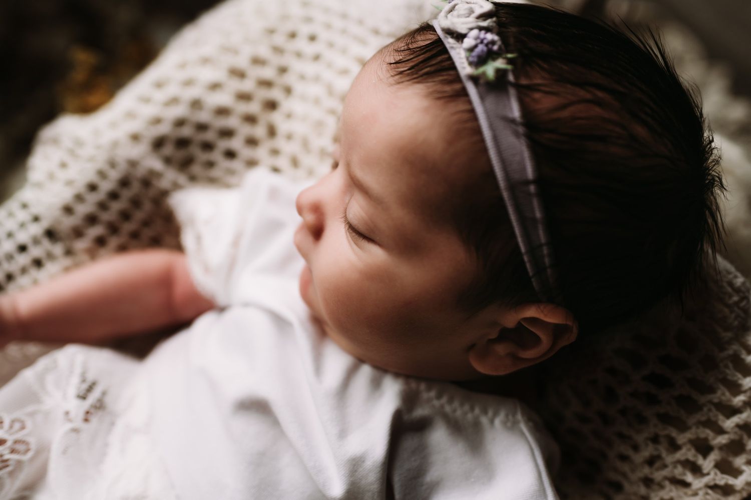 Baby wearing white dress and delicate headband resting on lace blanket in closeup profile.