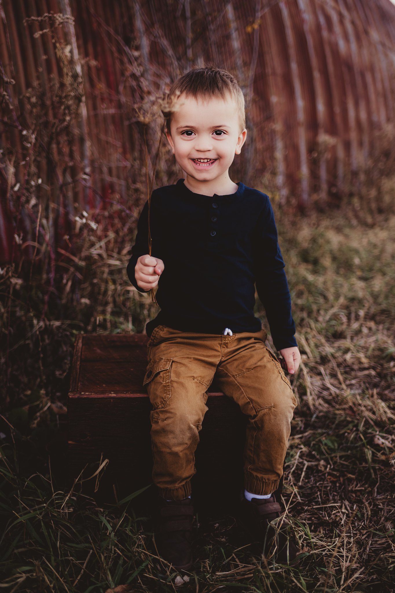 A smiling child in a black sweater and brown pants sitting on a wooden bench against a rustic barn wall.
