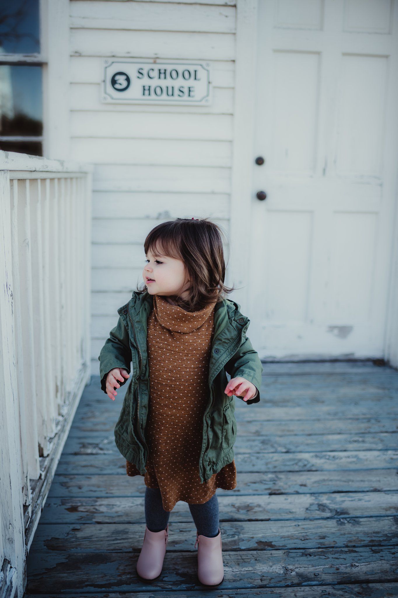 A young child in a green coat stands on a wooden porch outside a white schoolhouse.