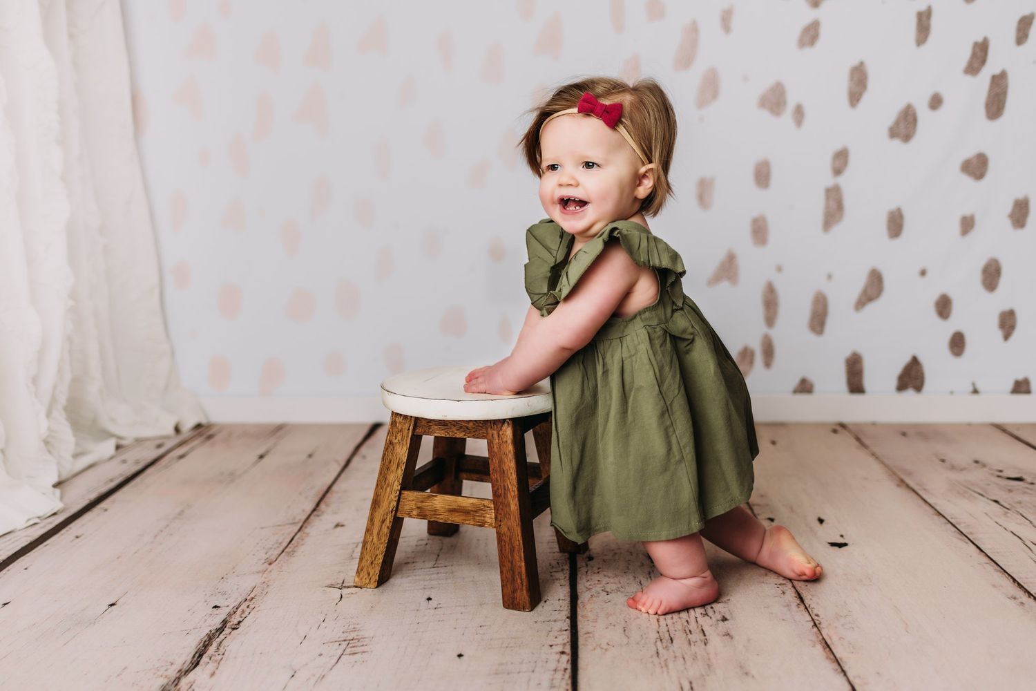 Joyful moment captured with wooden stool and olive green dress against patterned white background.
