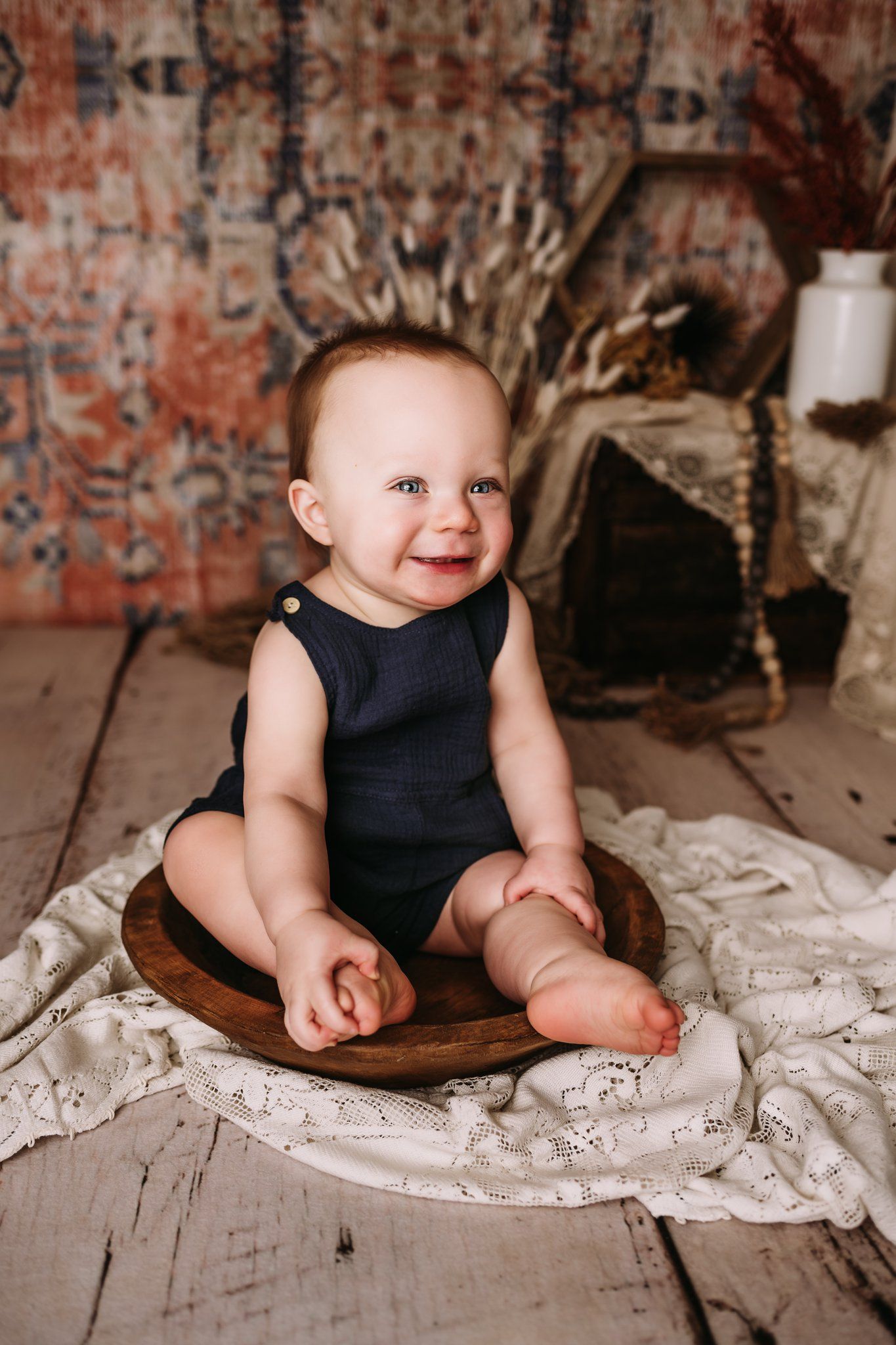Adorable baby in navy romper sits smiling on wooden bowl against vintage brick backdrop with lace blanket.