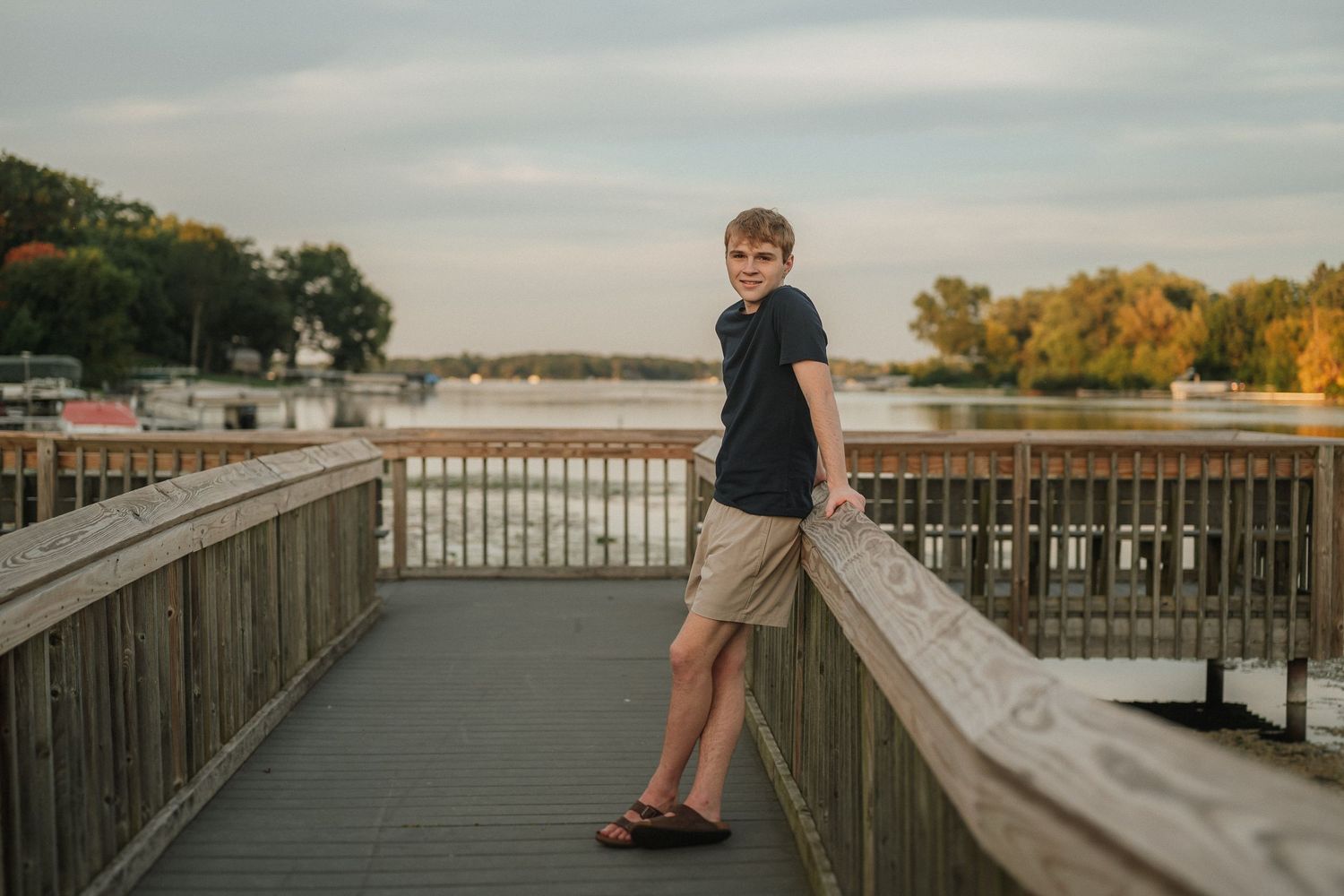 Person stands on wooden boardwalk overlooking water during sunset with cloudy sky.