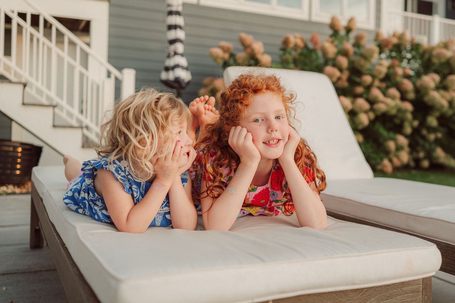 Two children with curly hair laying on a patio lounge chair share secrets and giggles on a summer day.