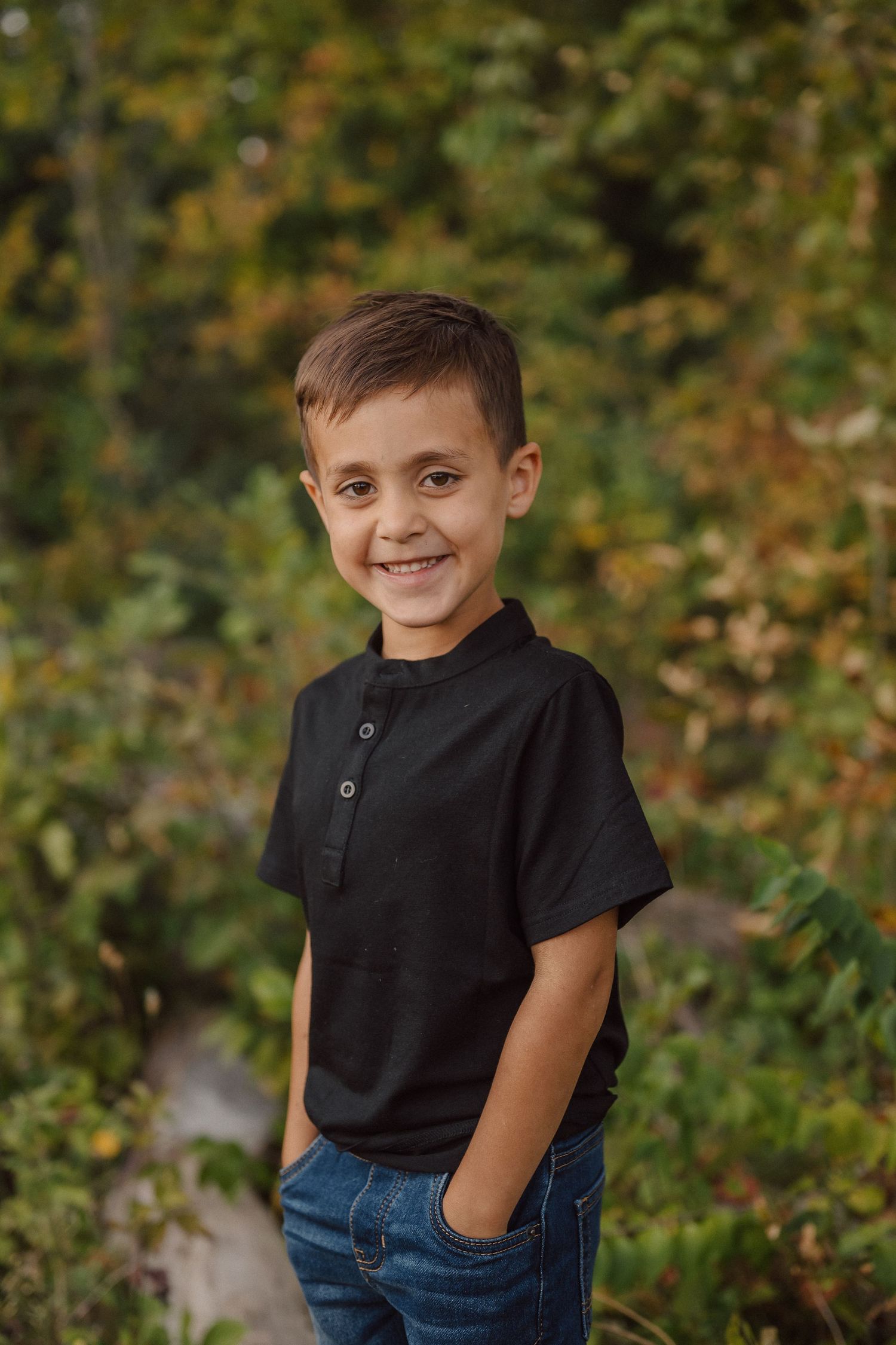A young child in a black polo shirt and jeans poses outdoors against a natural green background.