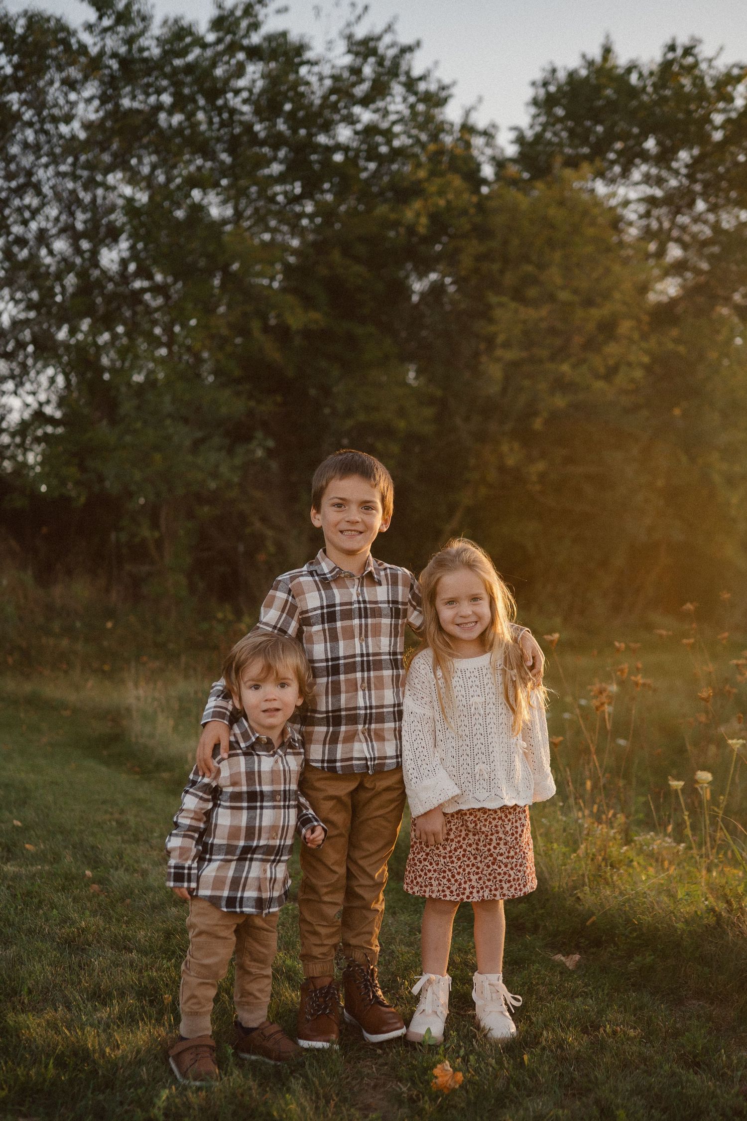 Three siblings pose together in a grassy field at sunset wearing coordinated casual outfits.