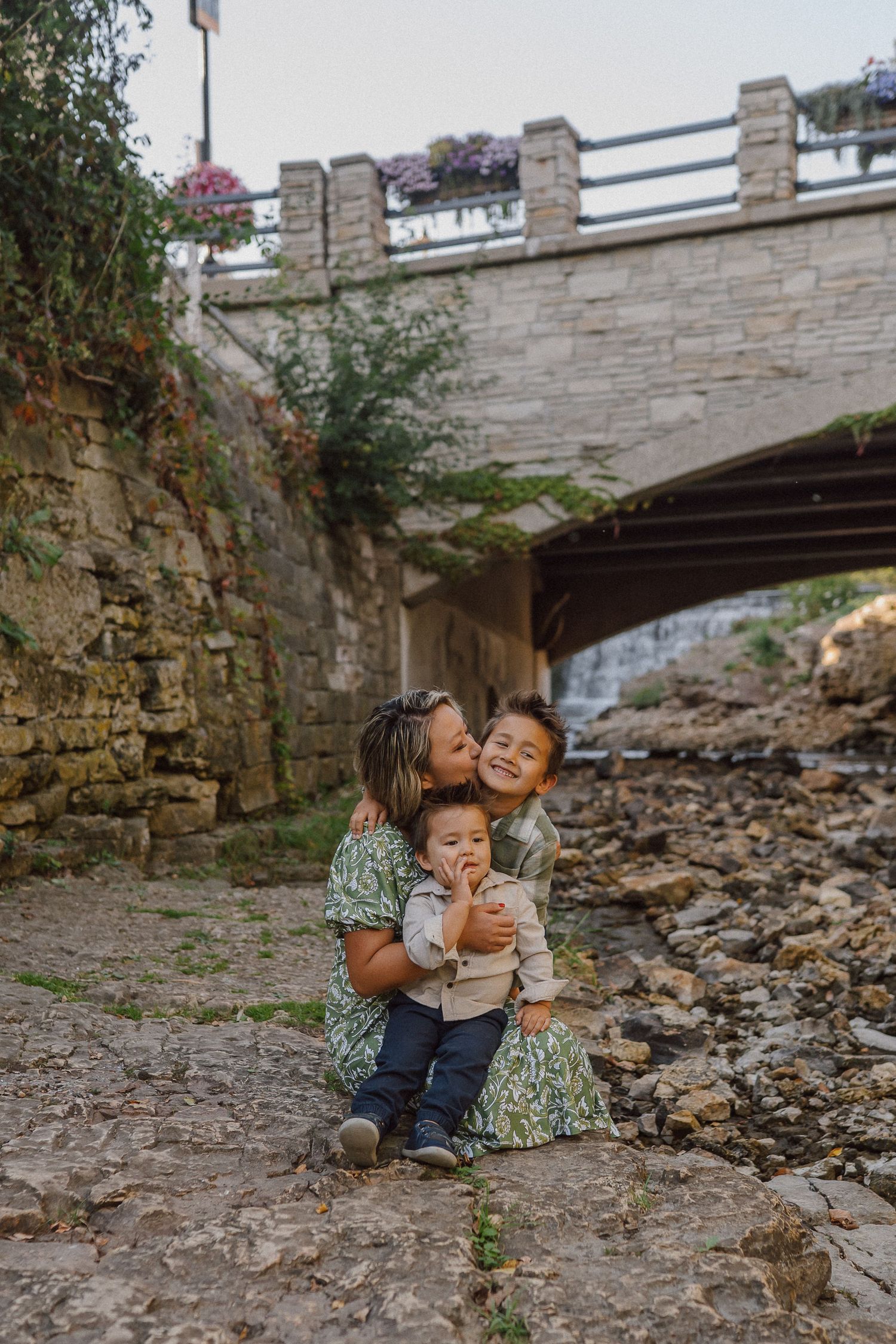 A sweet family moment captured under a stone bridge near a rocky creek bed.
