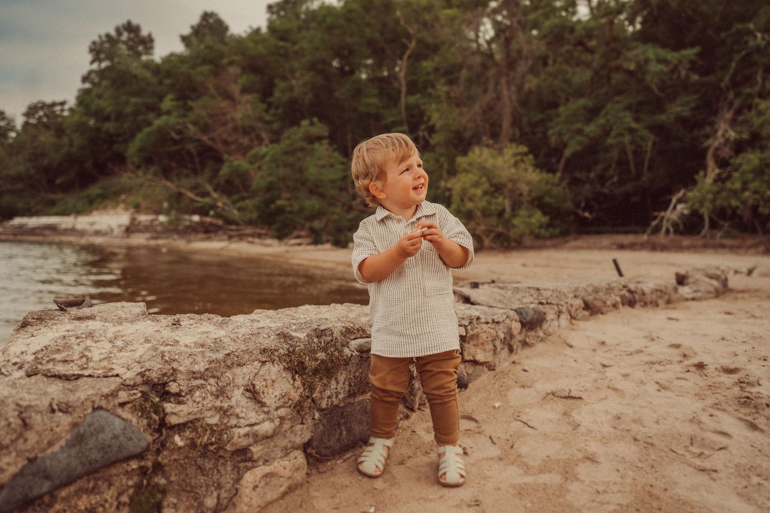 A toddler explores a sandy beach area near trees during golden hour.