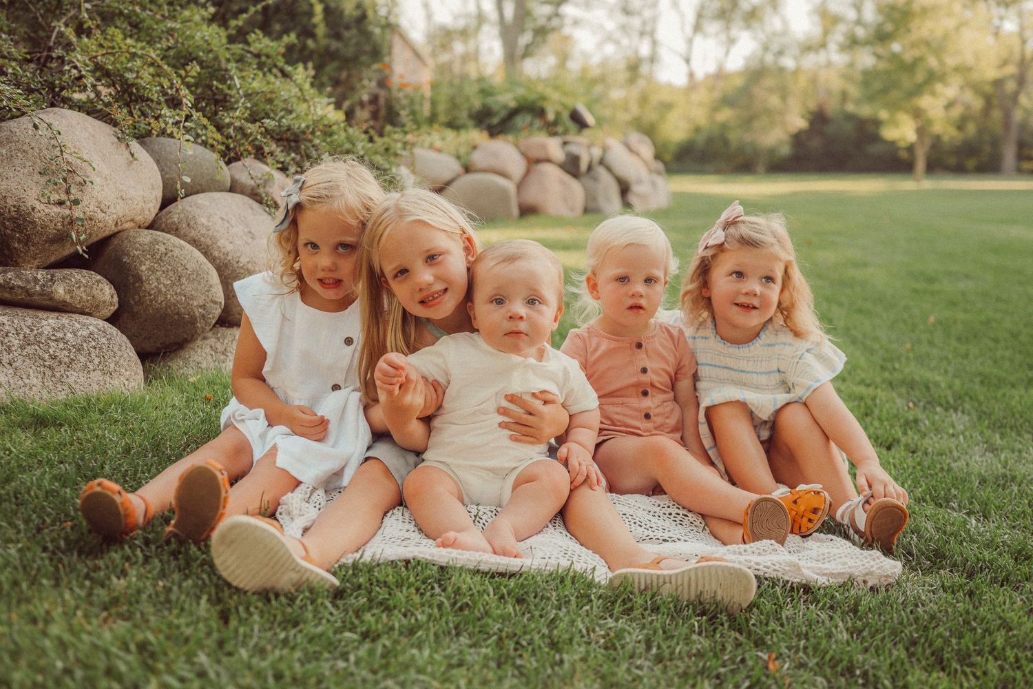 Children in coordinating white and beige outfits sitting together by a rock wall on grass.