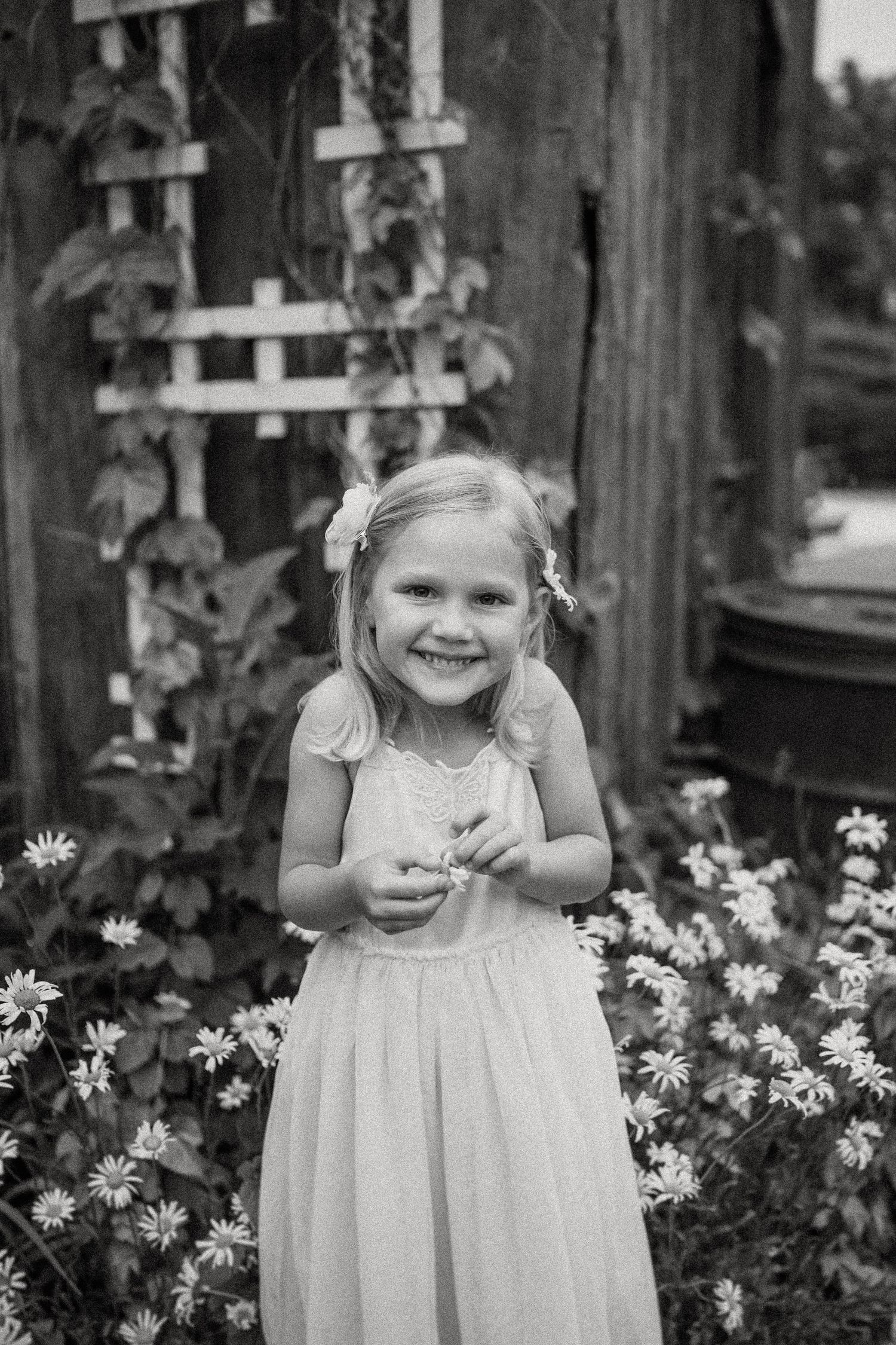 Black and white photo of joyful moment in flower garden beside rustic barn wall.