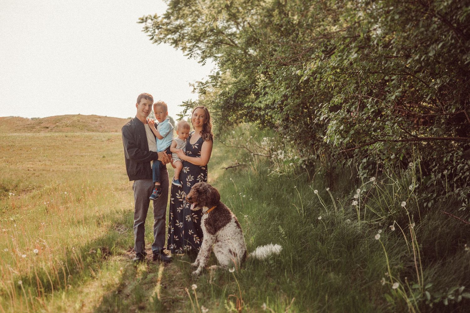 Family portrait in golden sunlight with their spotted dog on a grassy hillside.