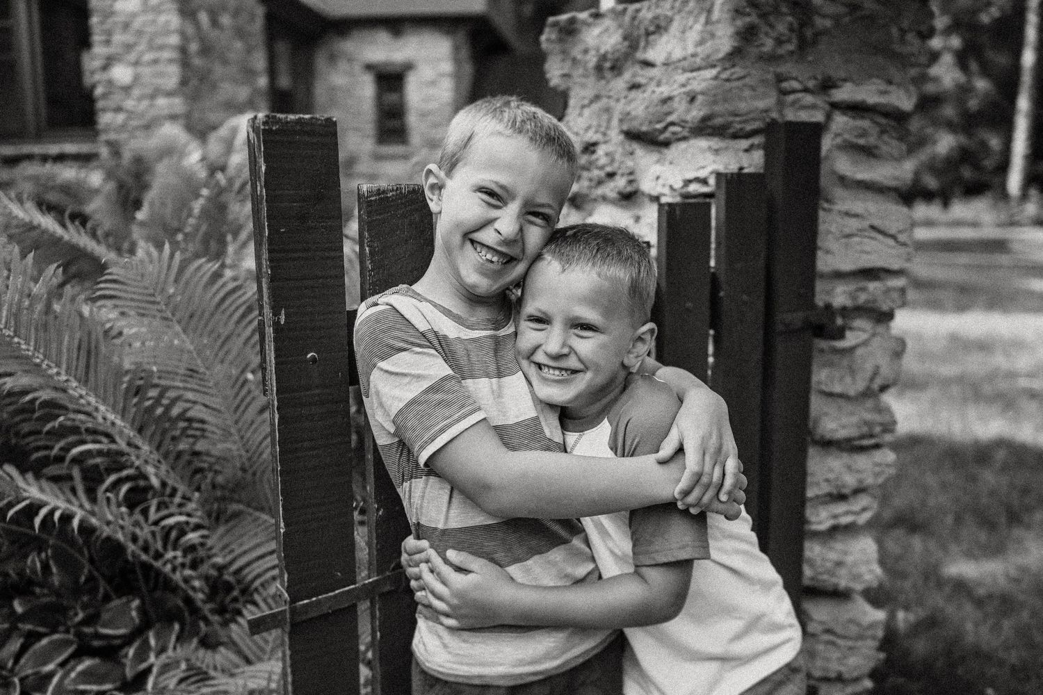 Black and white portrait of two boys sharing brotherly hug by garden gate.
