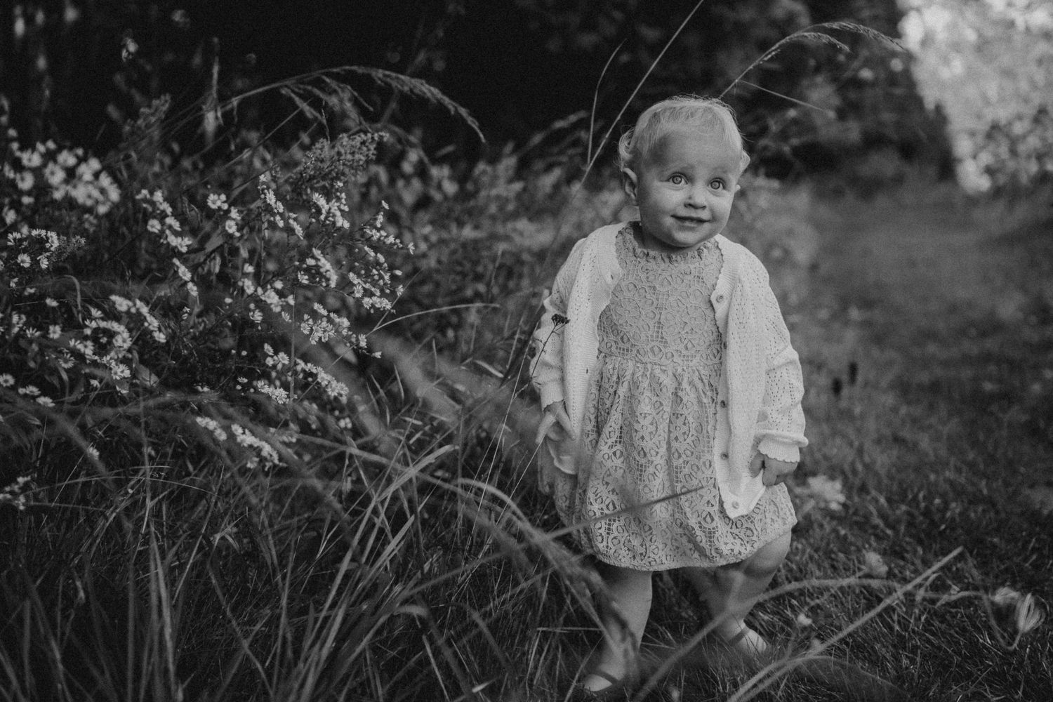 Black and white portrait of a child in a dress standing among tall grass and wildflowers.