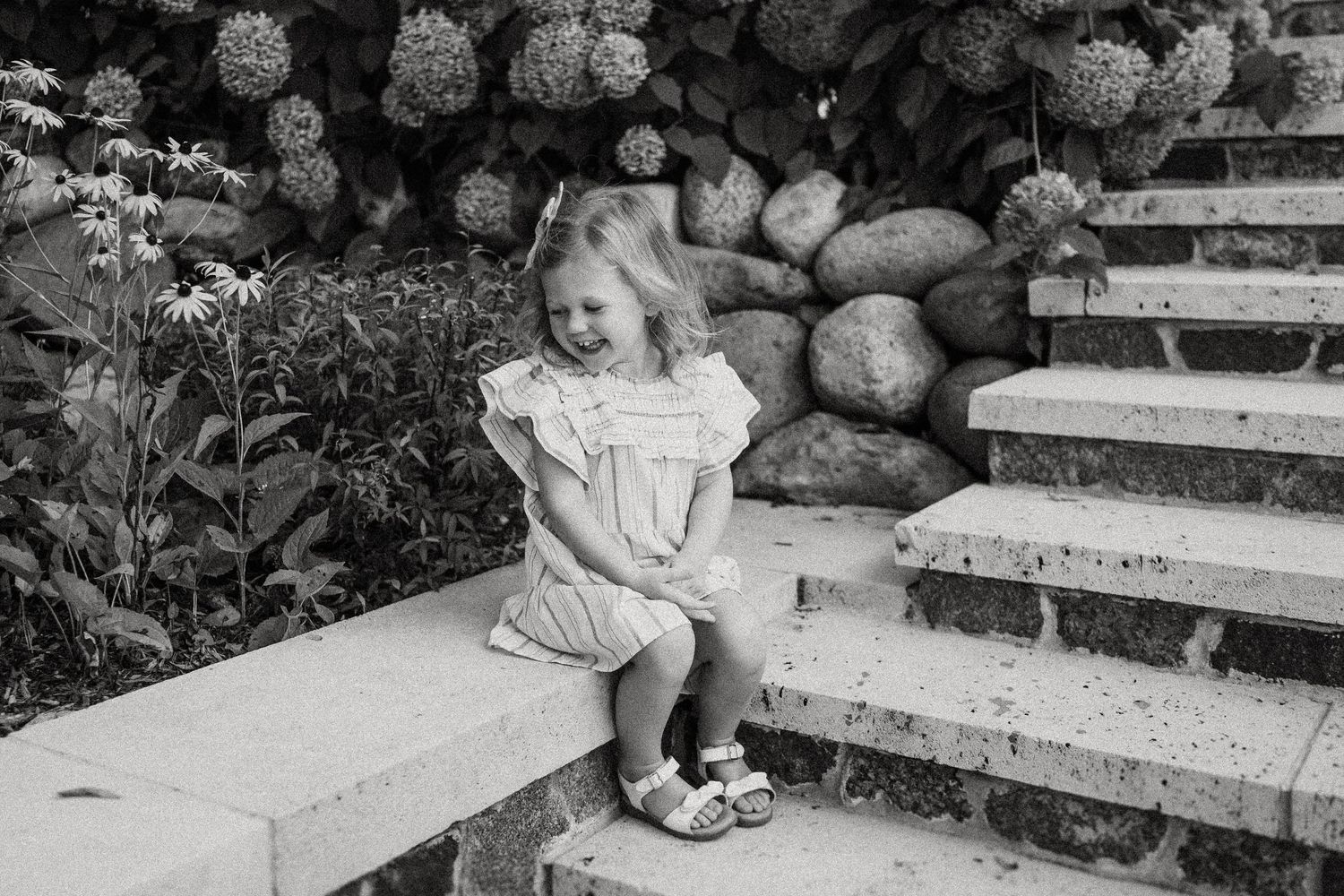Black and white photo of a child in a dress sitting on concrete steps near flowering shrubs.