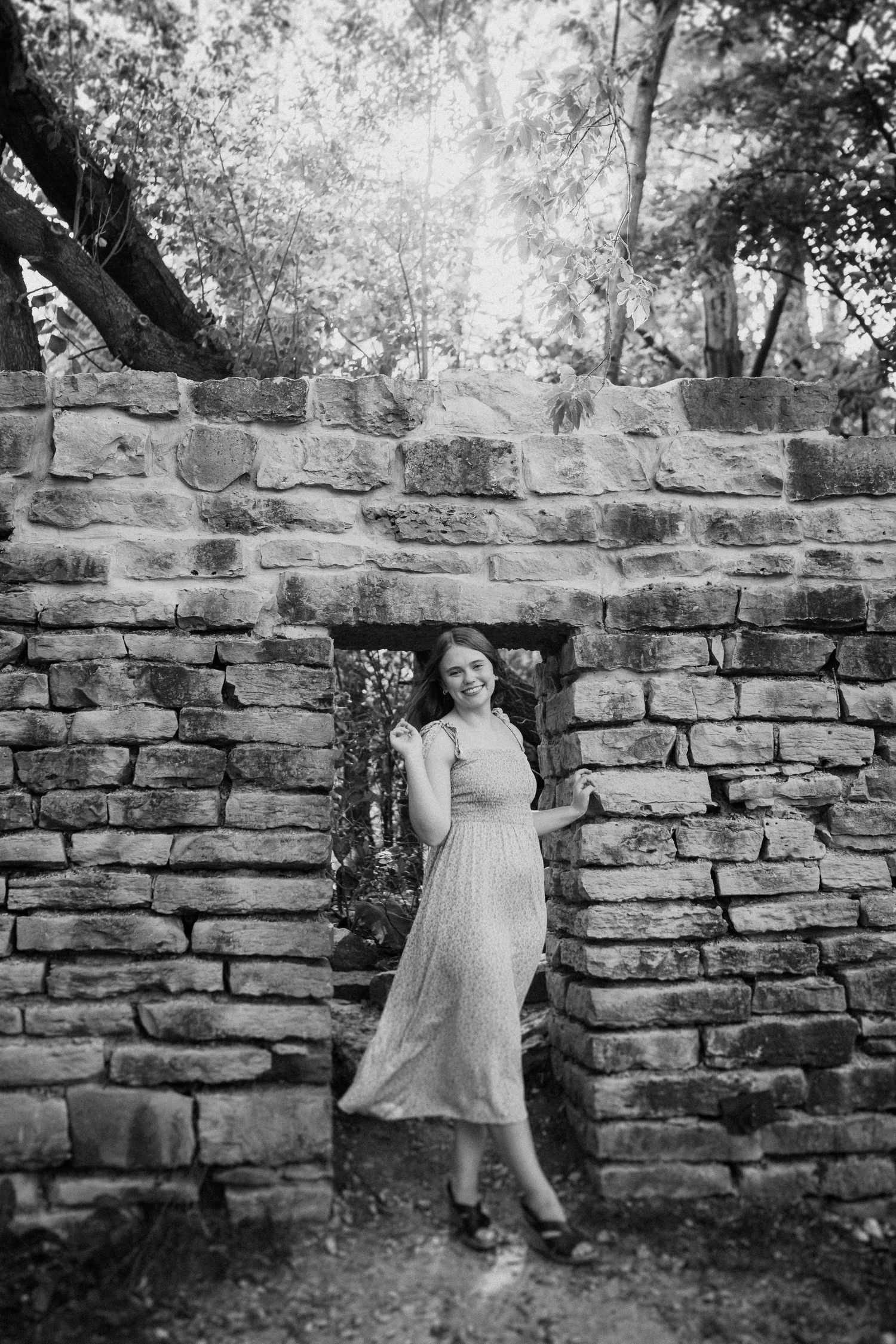 Black and white portrait of a woman in a long dress leaning against an old stone wall surrounded by trees.