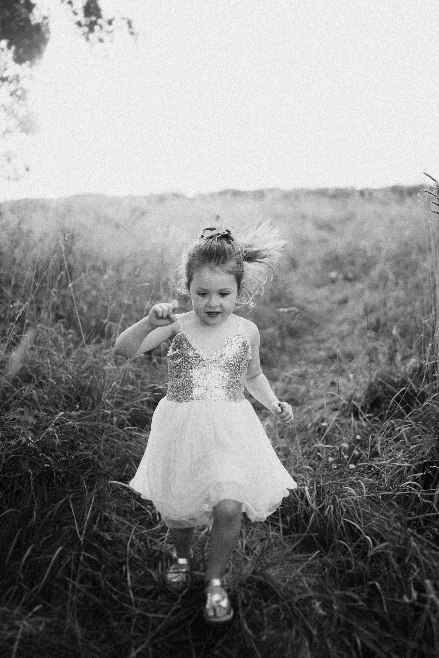 A young figure in a sparkly white dress dances through a field of tall grass in black and white.