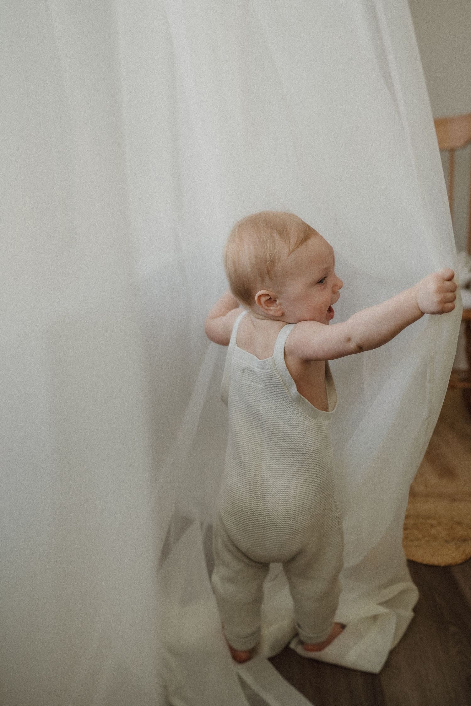 A baby in white overalls reaches for sheer curtains in a minimalist room setting.