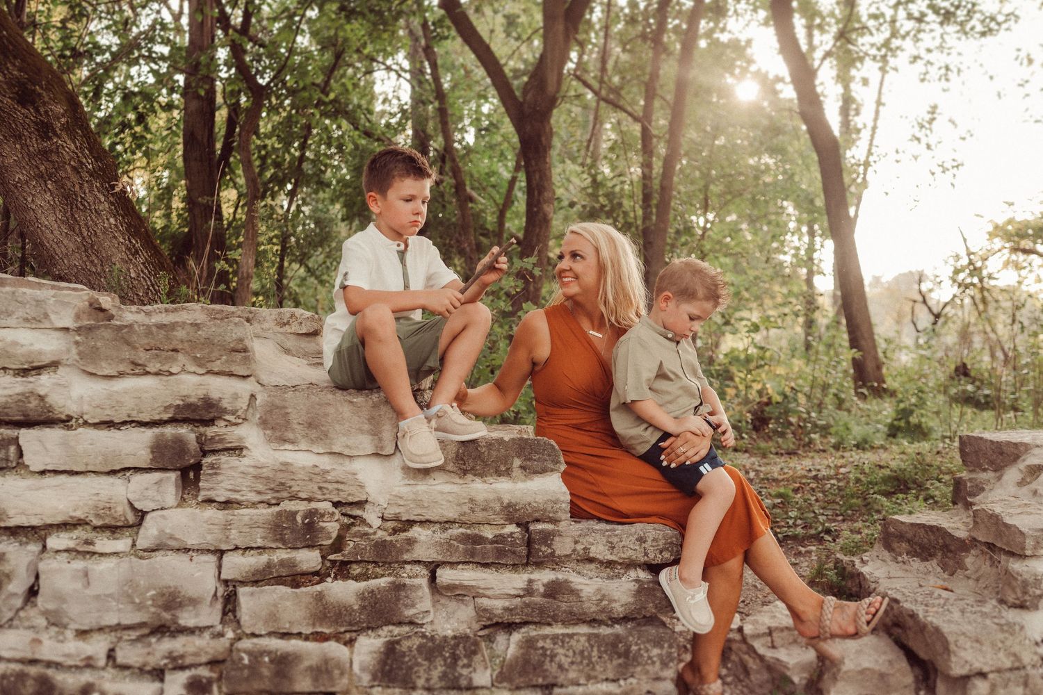 A family relaxes on rustic stone steps surrounded by sunlit trees in a wooded setting.