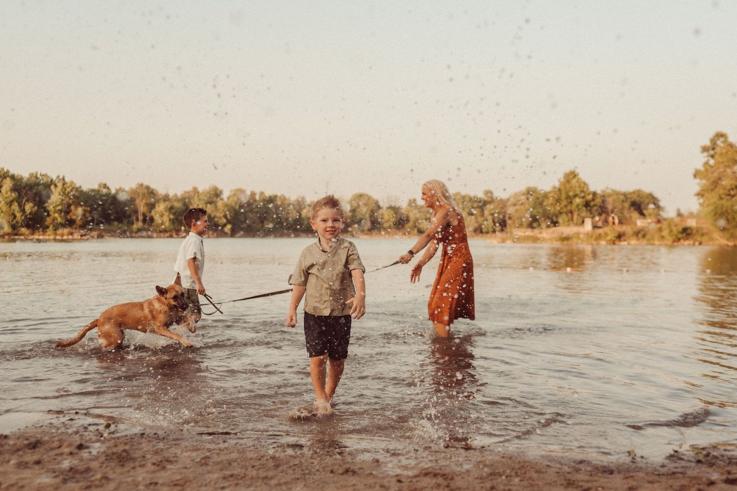 Family and dog enjoying a warm sunset while standing in shallow water near a tree-lined shore.