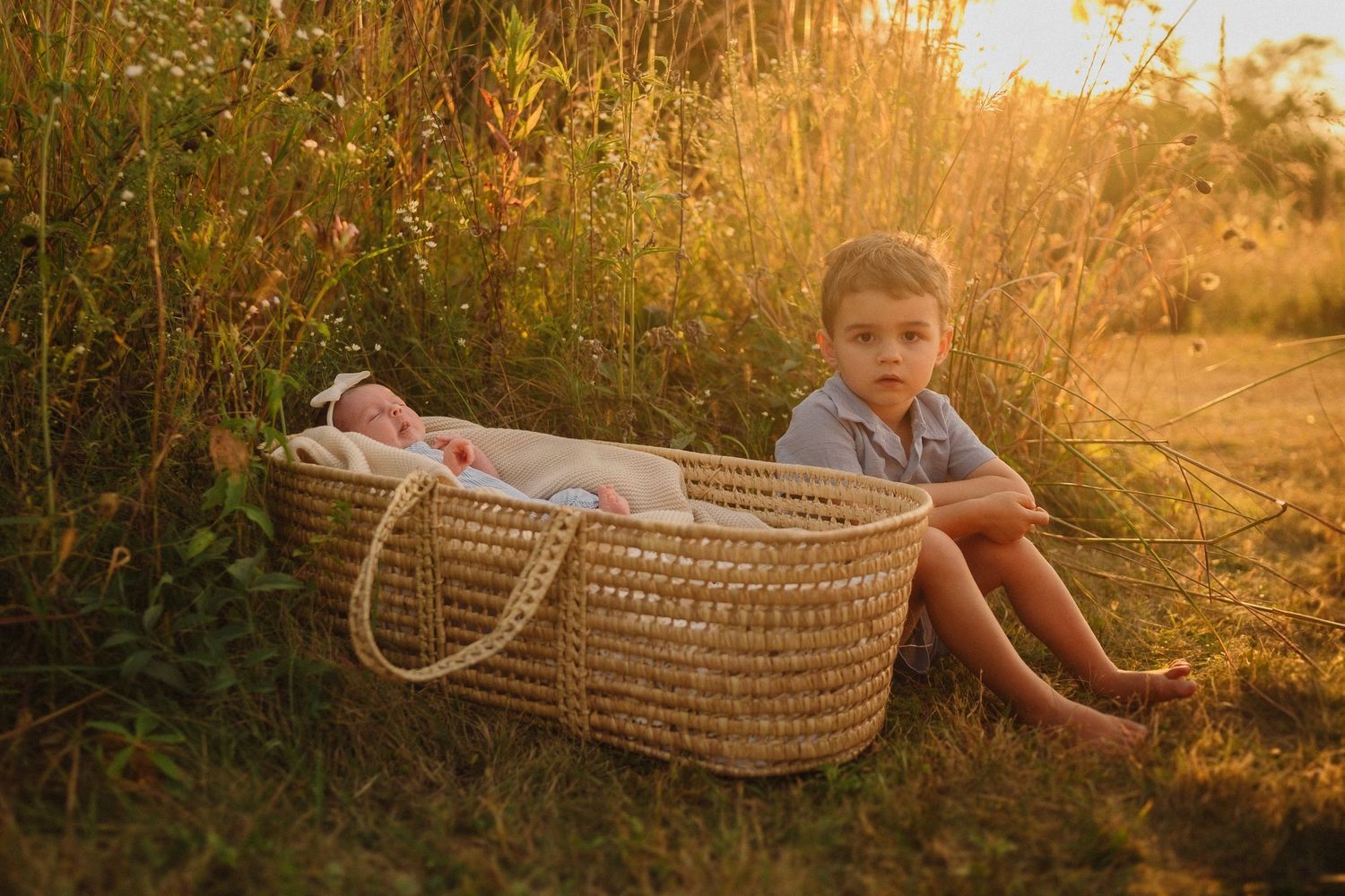 Child sitting beside wicker Moses basket in golden sunset field with tall grasses.