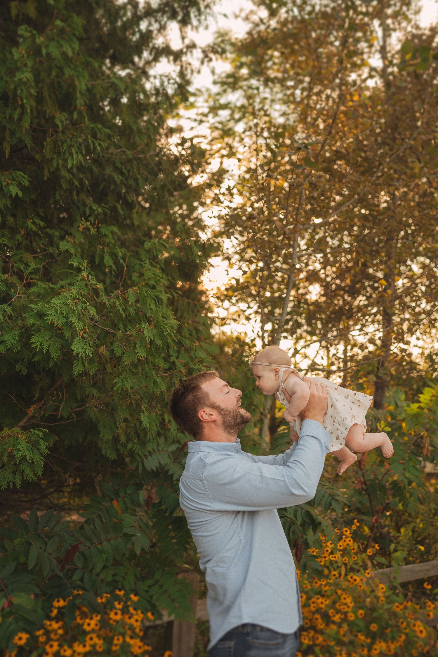A joyful moment as a parent lifts a smiling baby into the air against autumn trees.