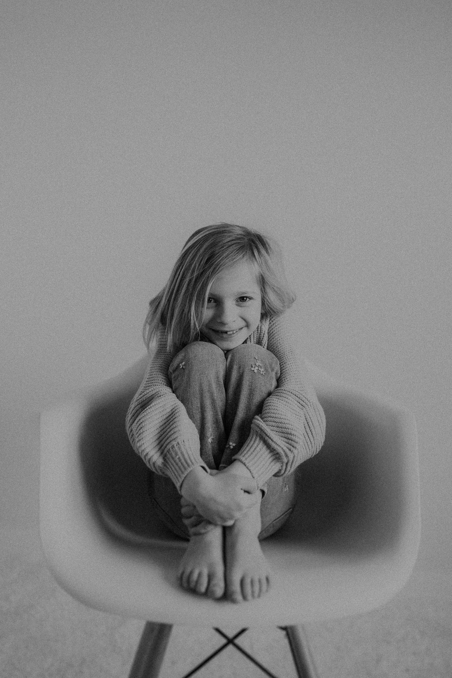 Child hugging knees while sitting on modern chair in casual black and white portrait.