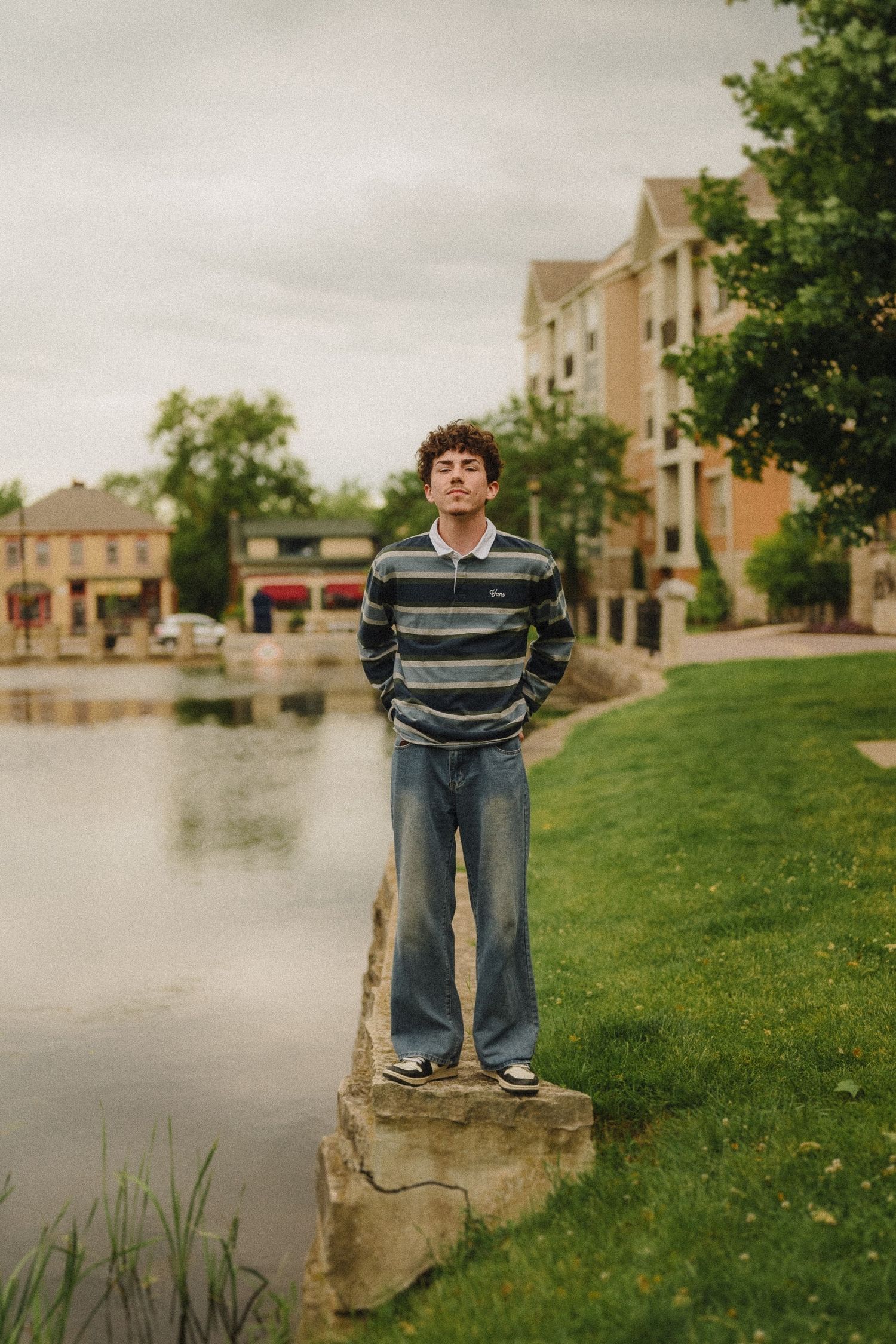 A person in a striped sweater and jeans stands on a concrete ledge beside a pond with buildings in the background.