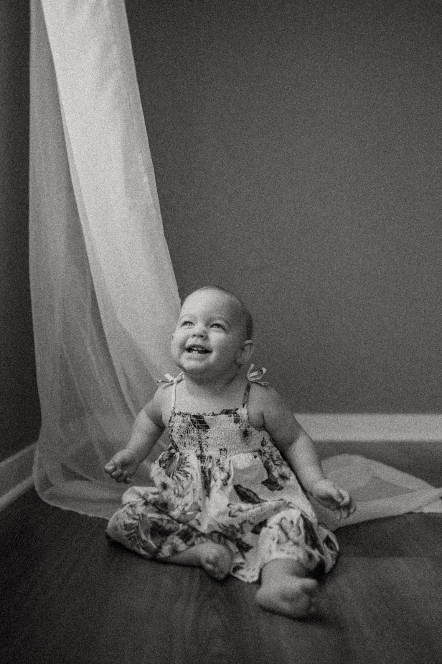 A black and white photo of a laughing toddler in a dress sitting by flowing curtains.
