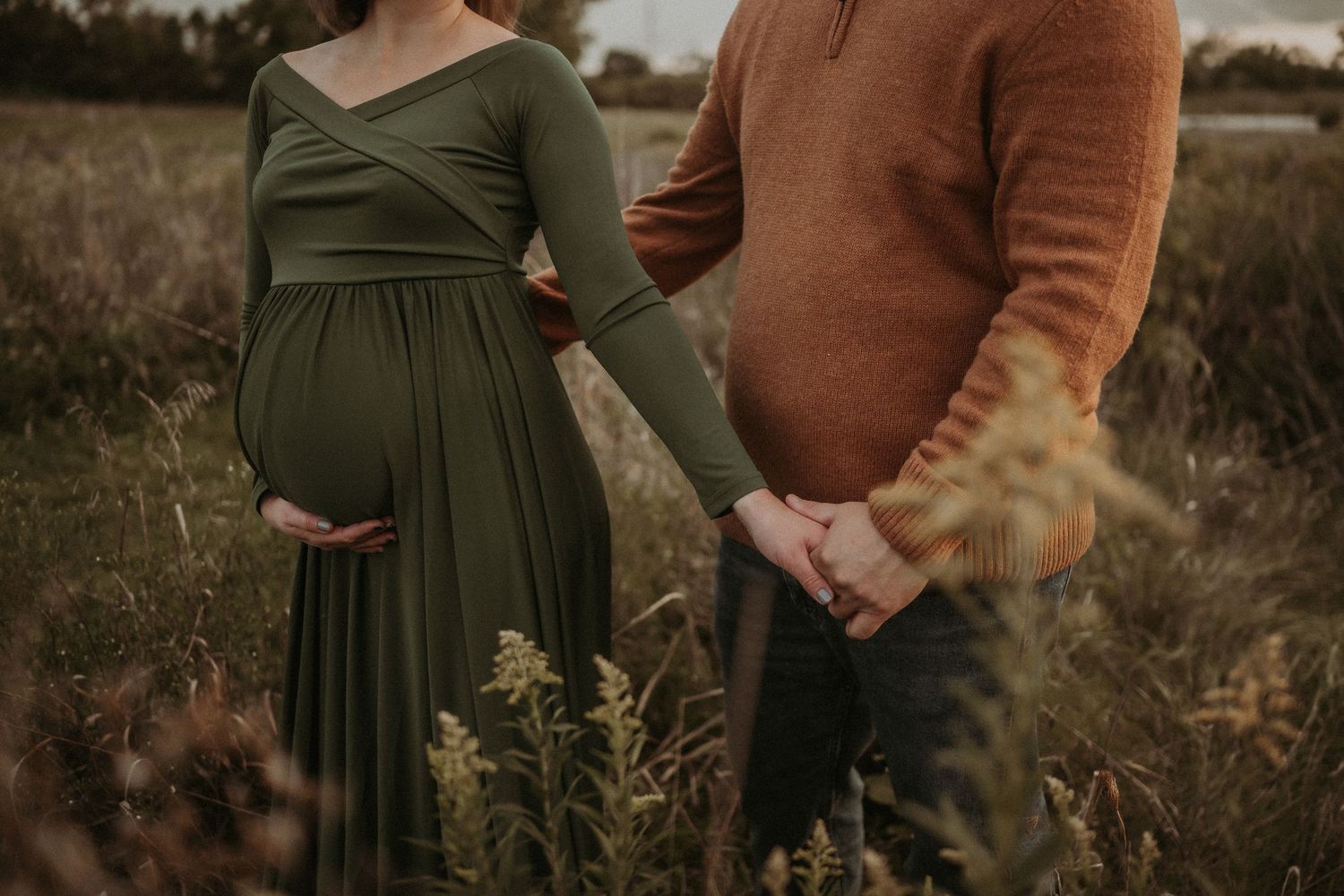 Side view of expectant couple holding hands in wild grass field at dusk.