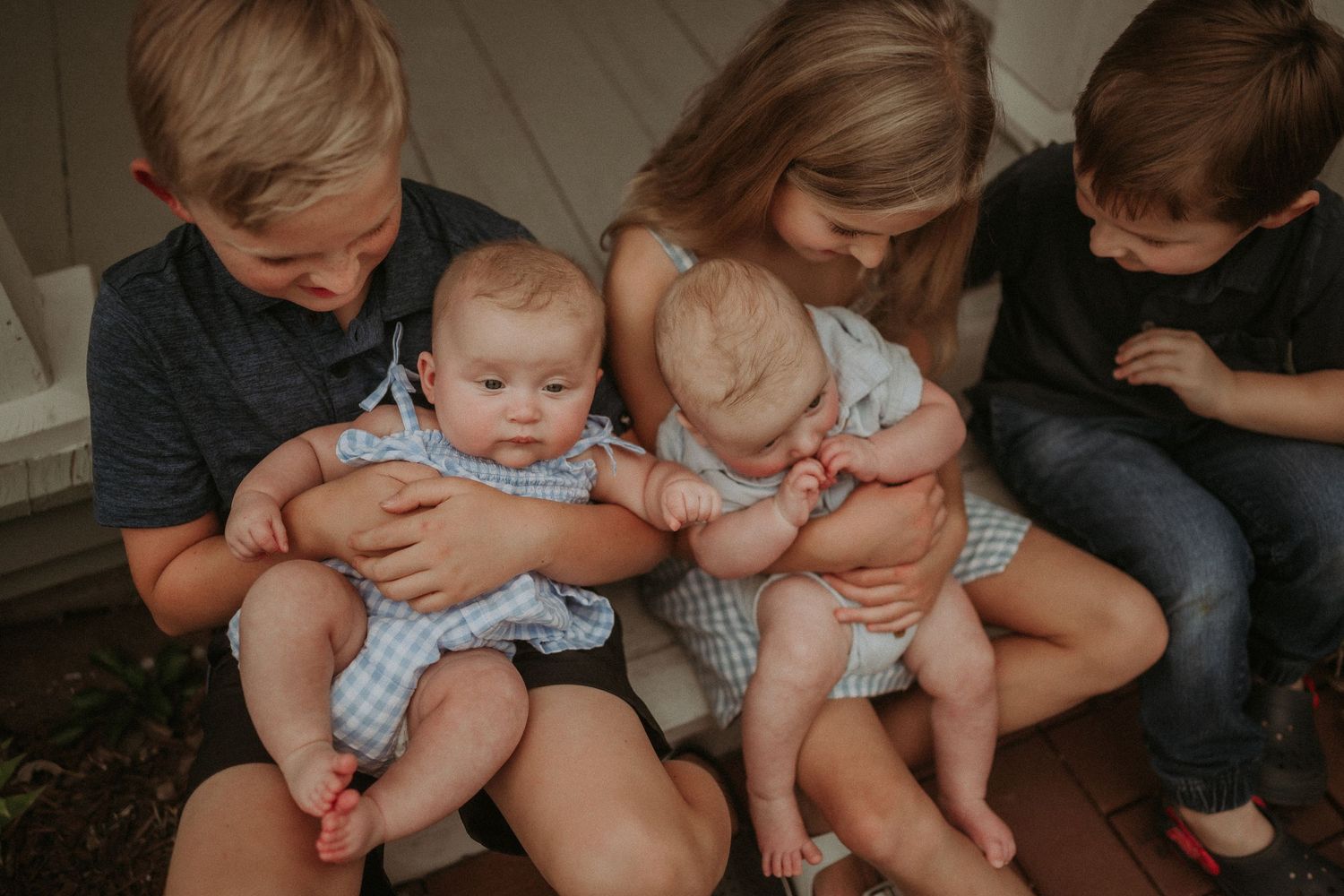 Siblings sitting close together on porch steps holding twin babies in matching outfits.