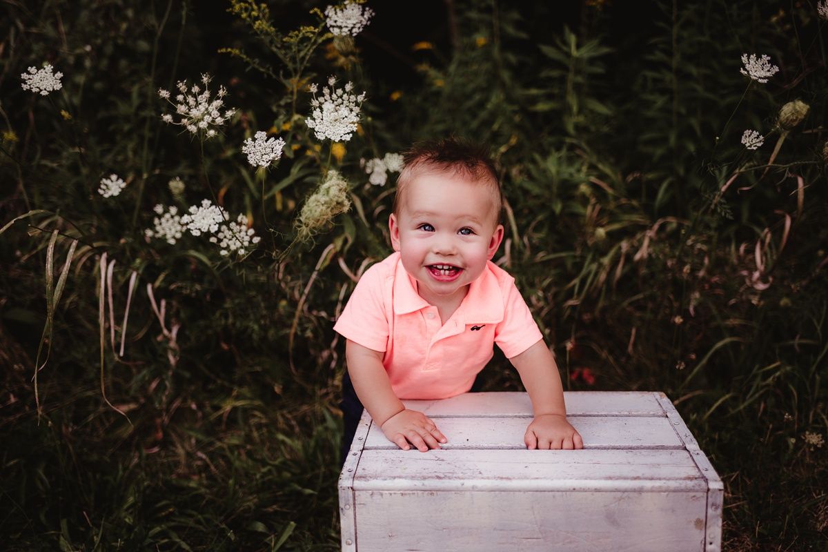 A baby in a pink shirt smiles while sitting in a white wooden box surrounded by white flowers in a garden.