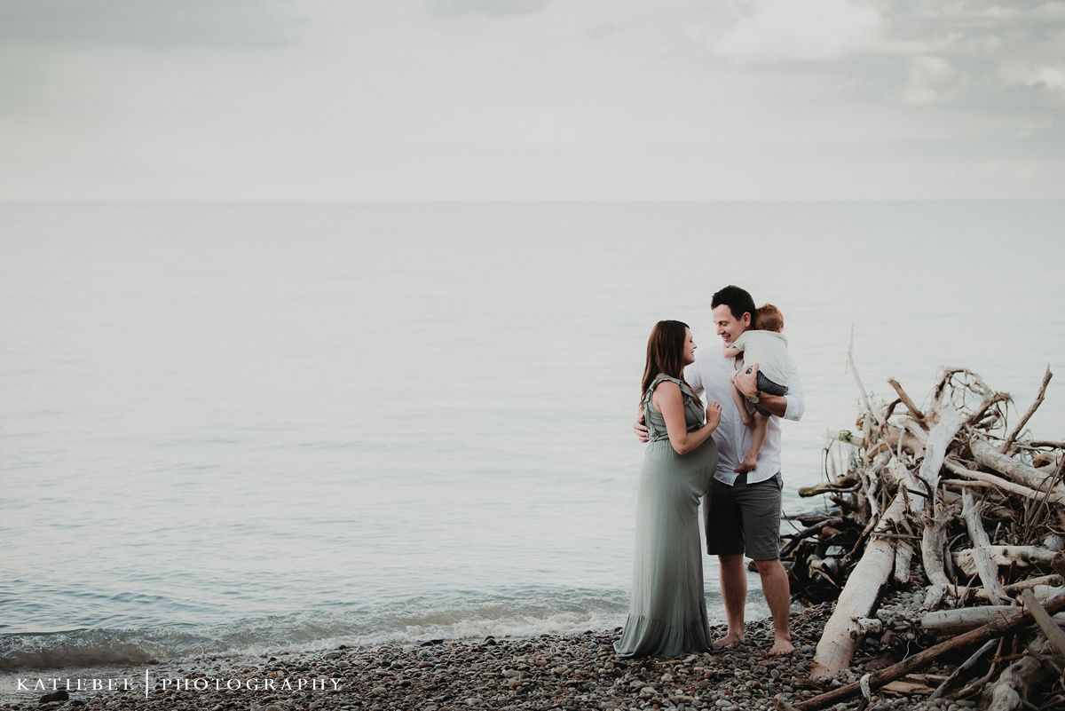 A couple embraces on a rocky beach with driftwood and misty ocean backdrop at sunset.