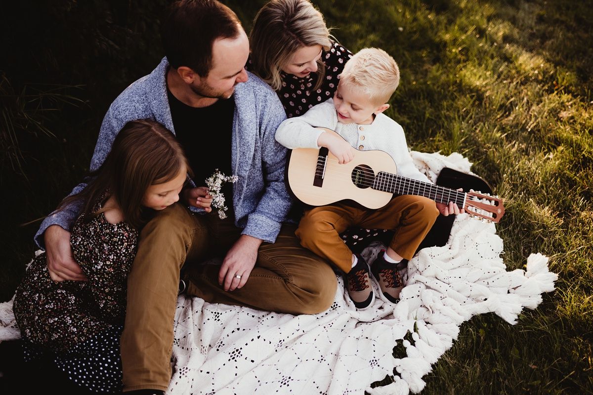 Family gathered on white blanket for outdoor music session with acoustic guitar.