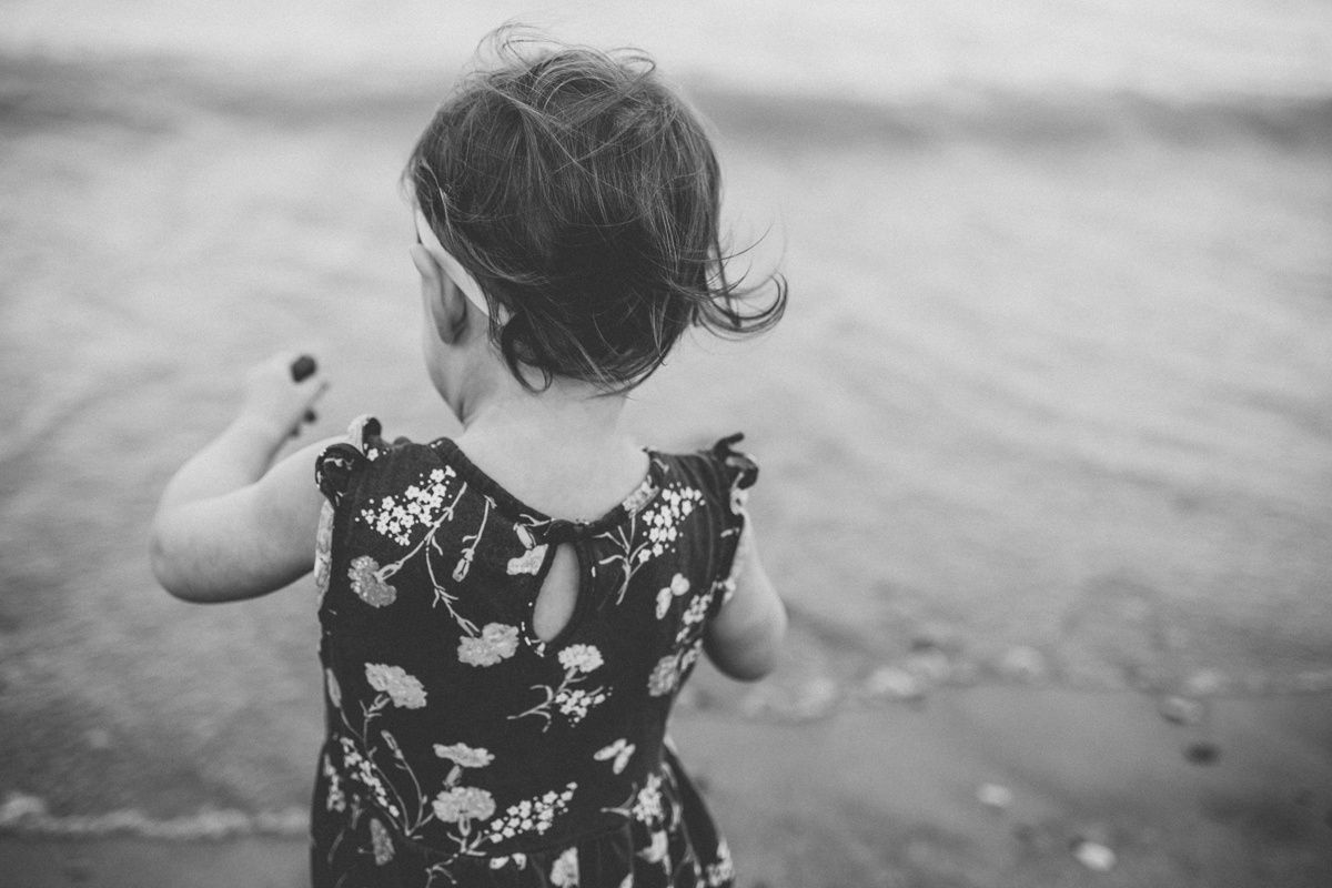 A small child in a floral dress looks out at the ocean waves in black and white.