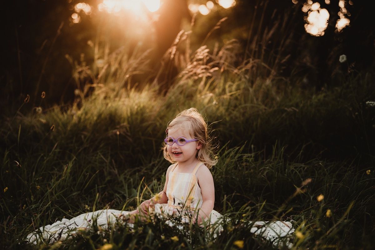 A child in a light dress sits among wildflowers during golden hour sunset.