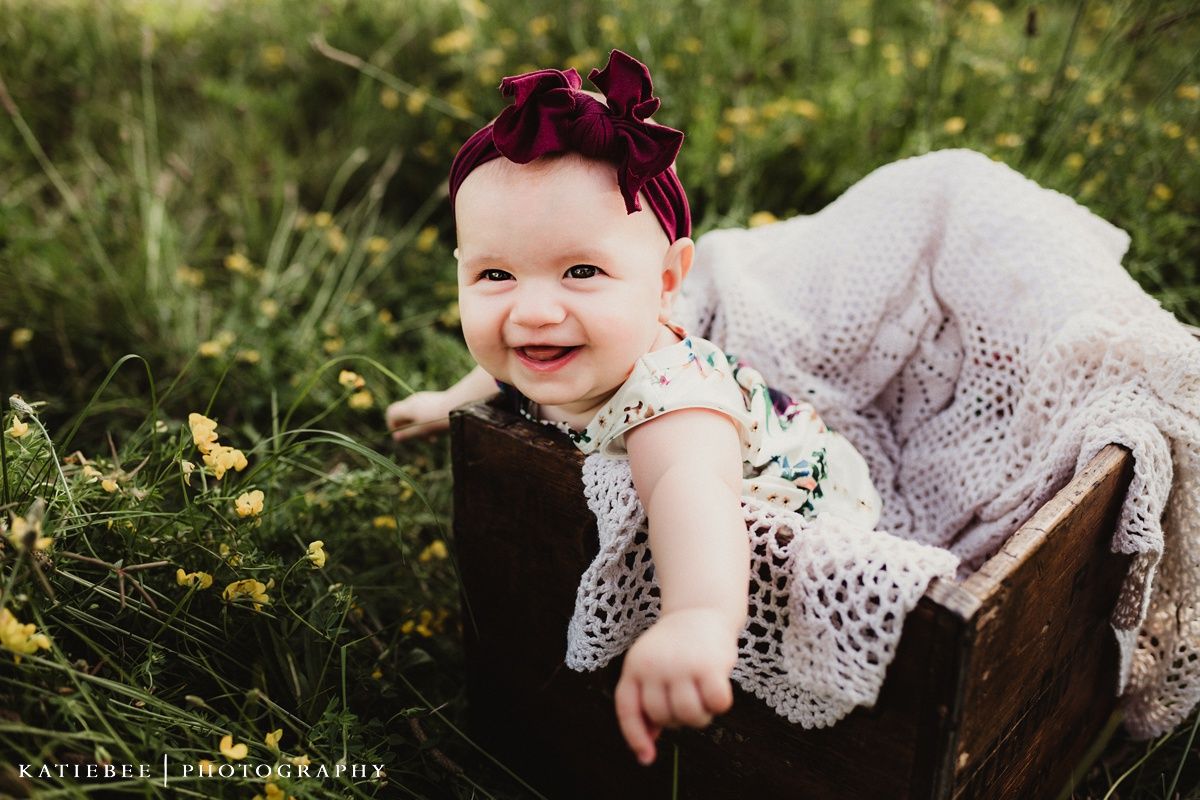 Baby in white lace dress and burgundy bow laughs while sitting in wooden box outdoors.