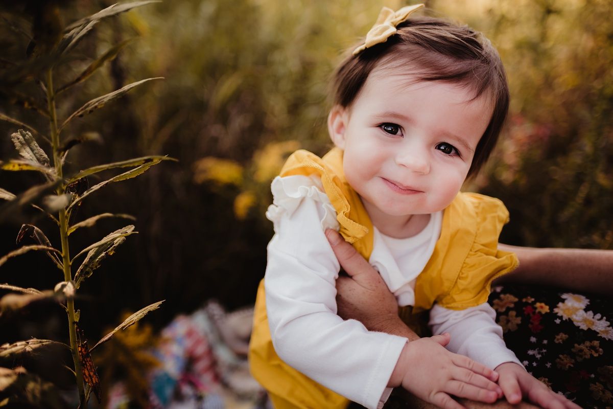 A toddler in yellow and white clothing smiles while playing outdoors.